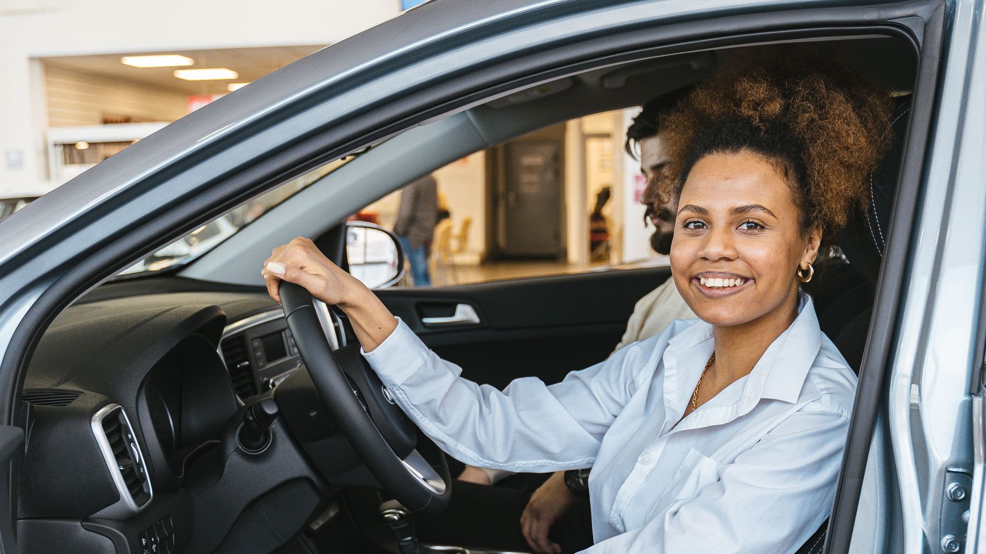Smiling woman sitting in a car at a dealership, ready for a test drive.