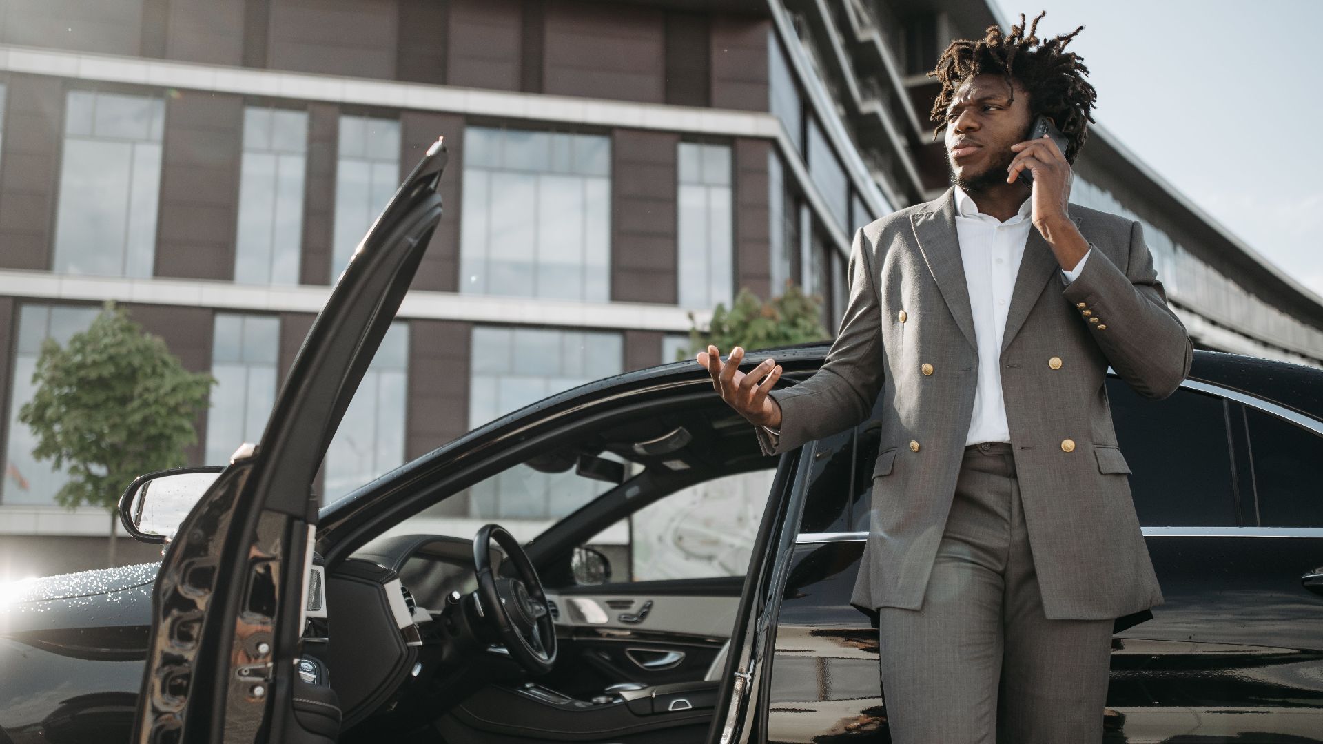 Confident businessman in a gray suit standing by a luxury car while talking on a smartphone.