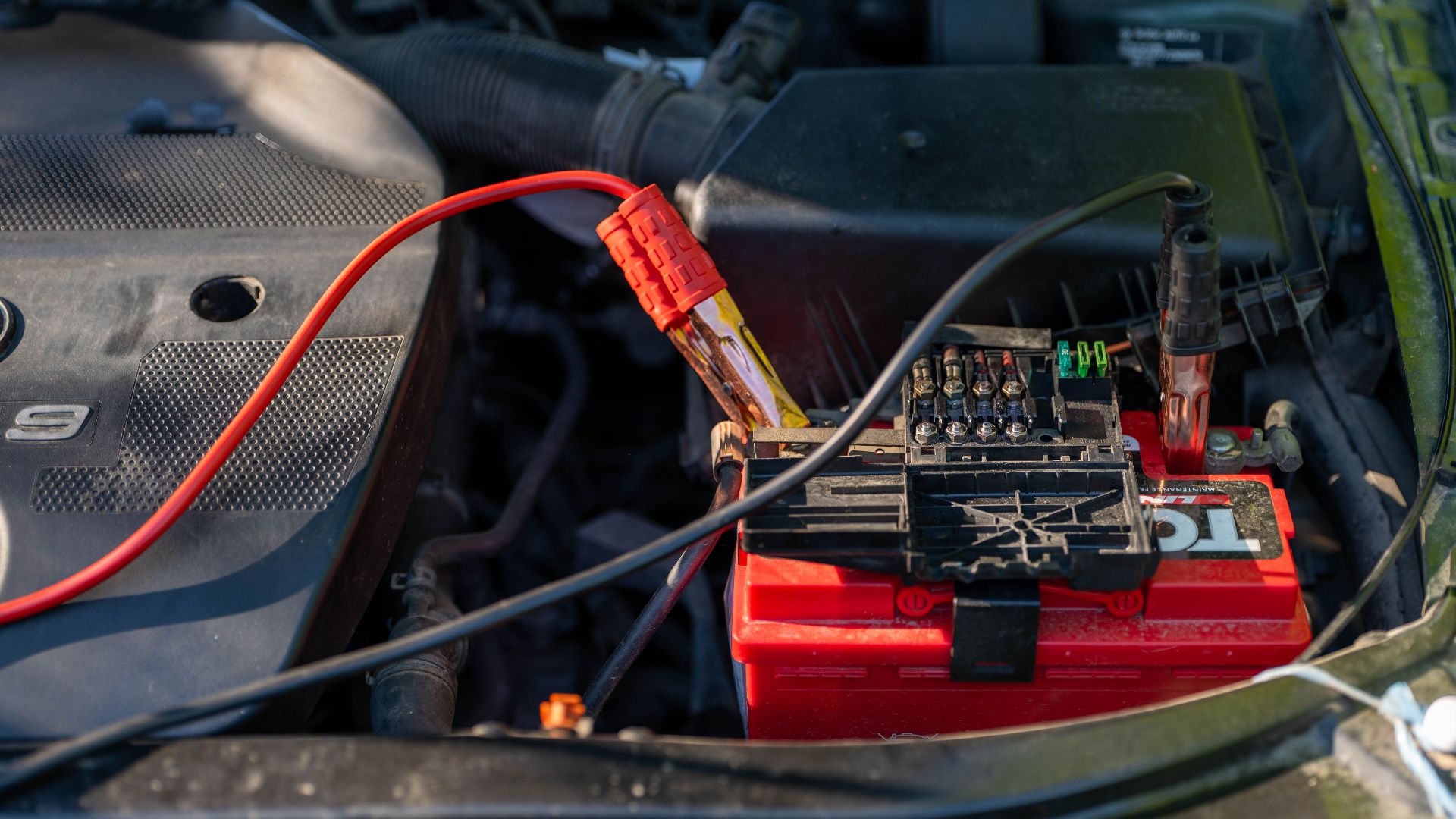 Close-up of a car battery with attached jumper cables in an engine bay.