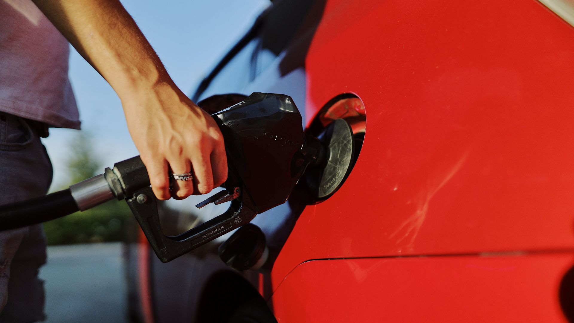 Close-up of a person refueling a red car at an outdoor gas station during the day.