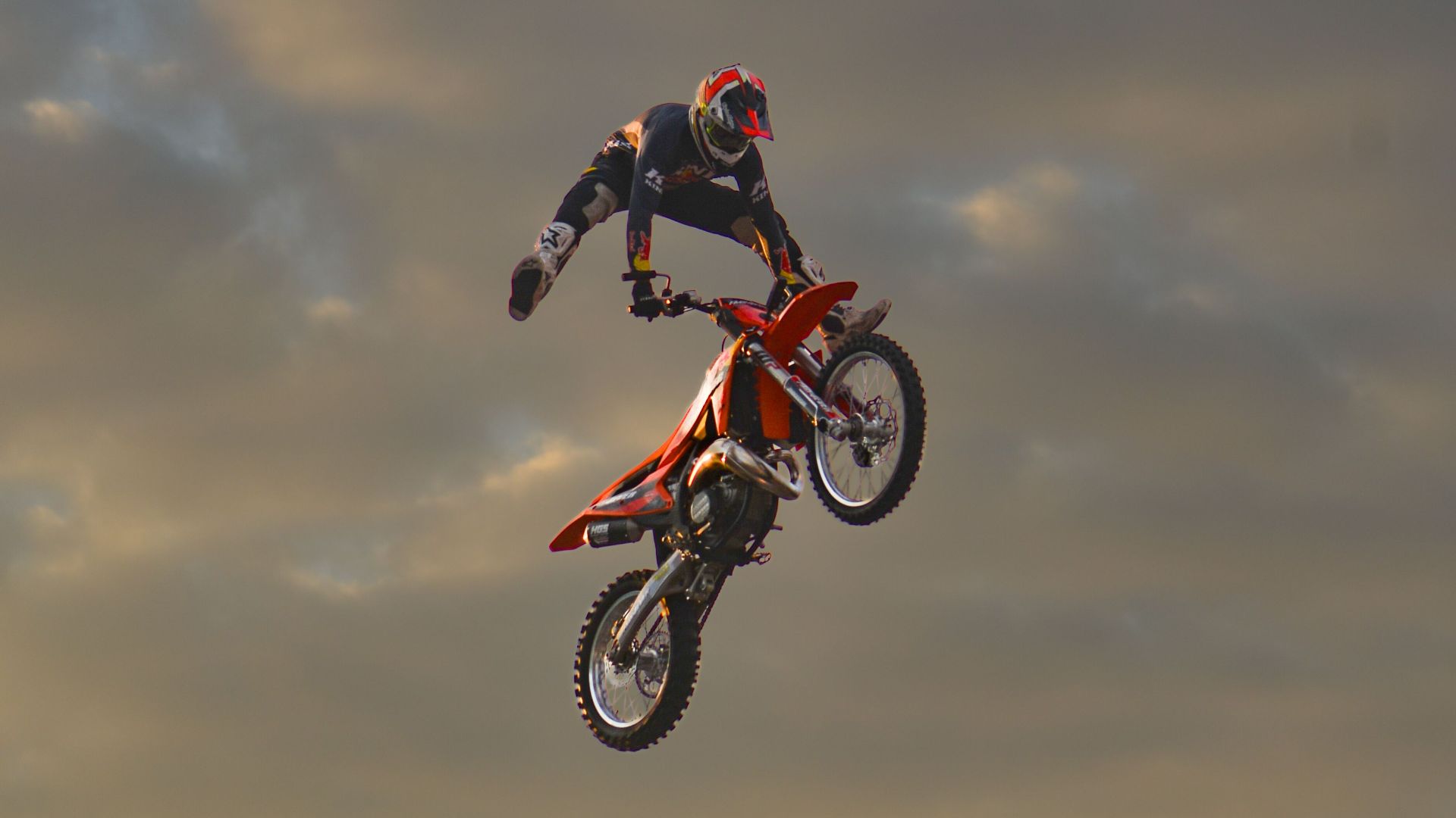 A motocross rider performs an aerial stunt against a dramatic cloudy sky during sunset.