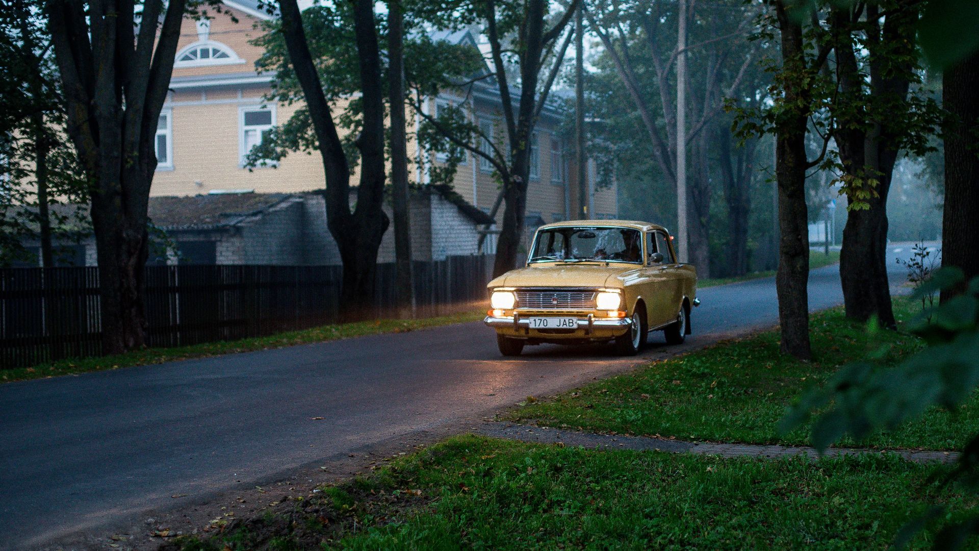 A classic yellow car driving on a quiet road lined with tall trees in a serene neighborhood.