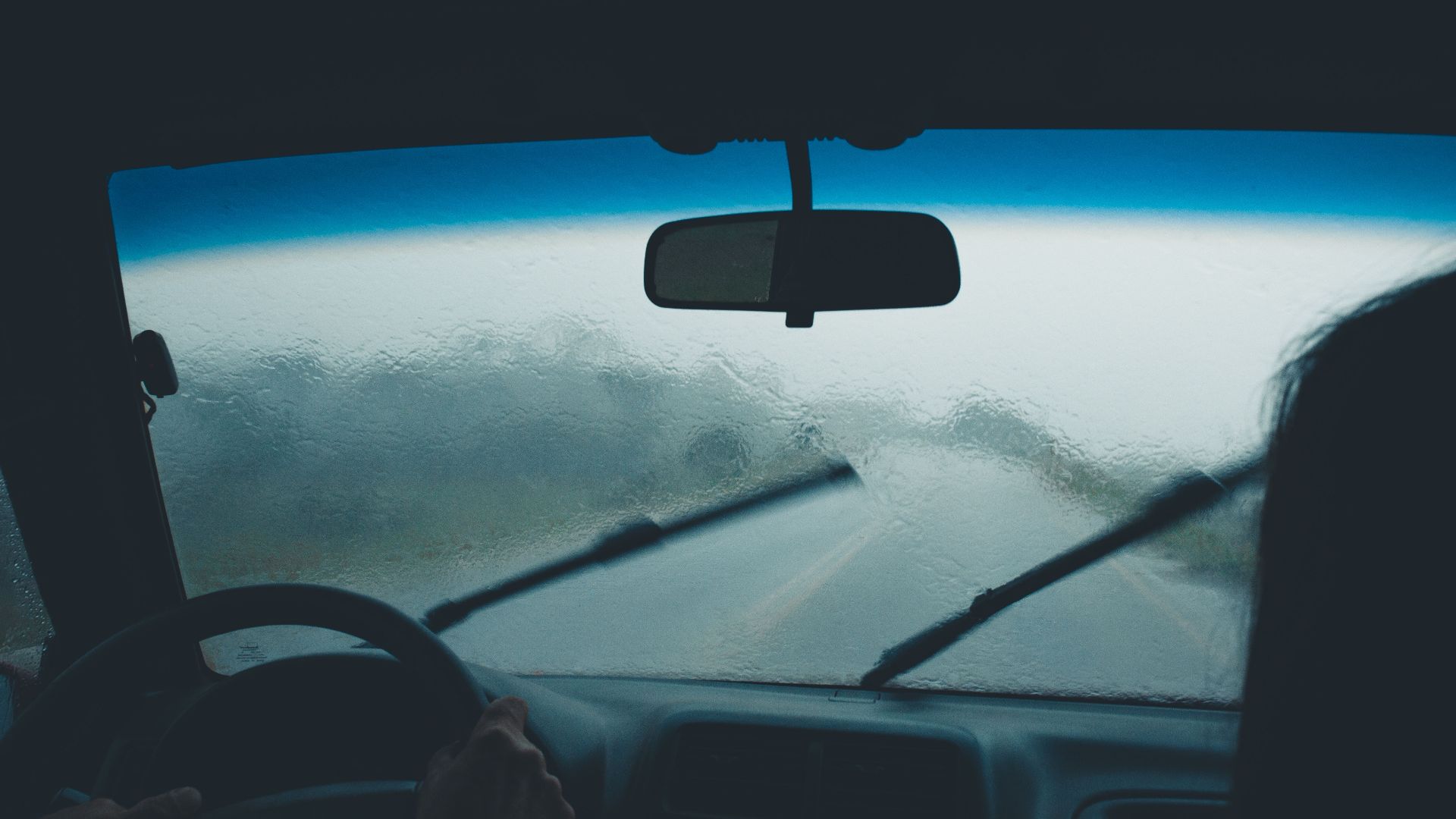 Interior car view on a rainy day with water droplets on windshield and wipers in motion.