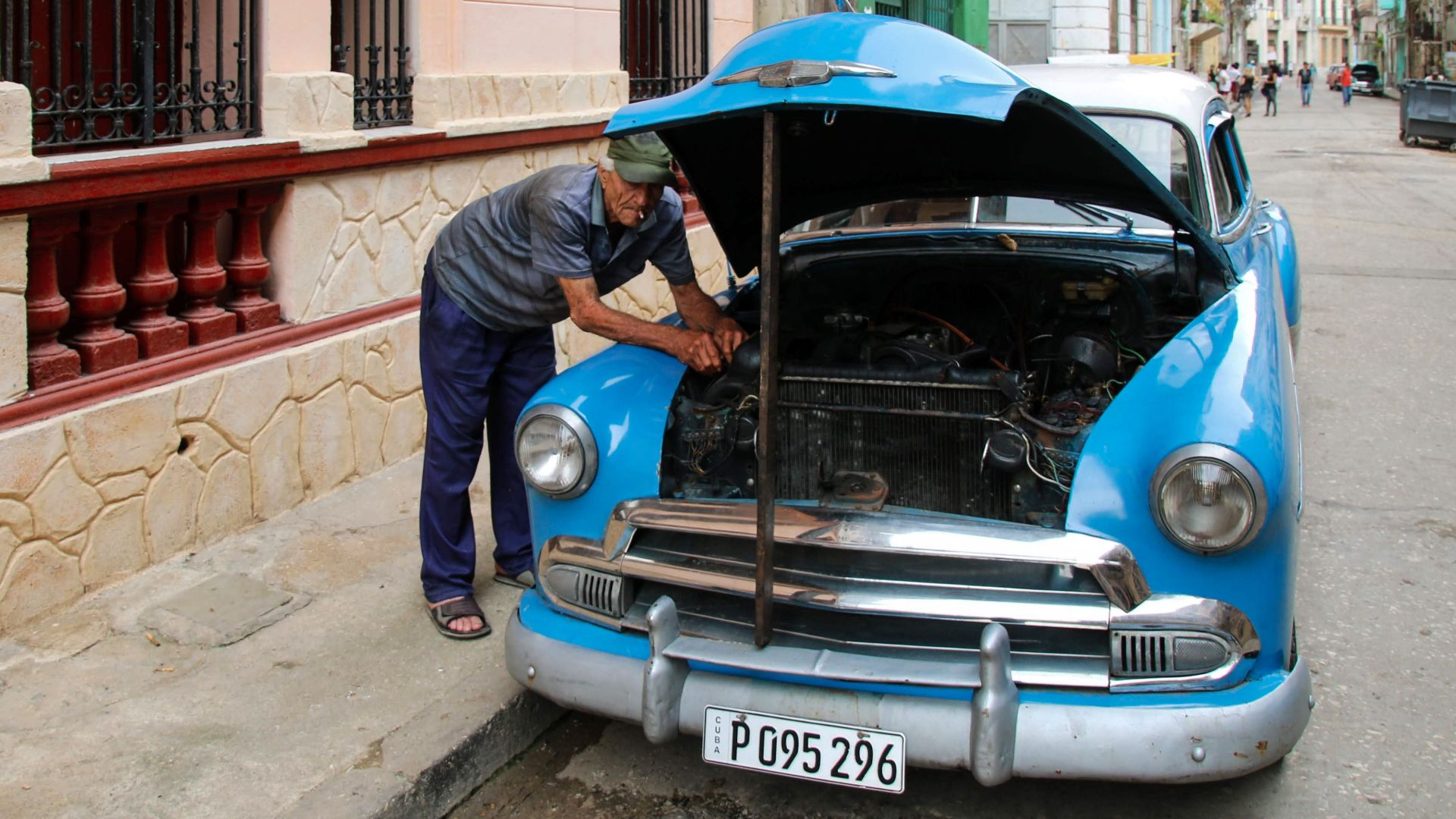 A man working on a vintage blue car's engine parked along an urban street with classic architecture.