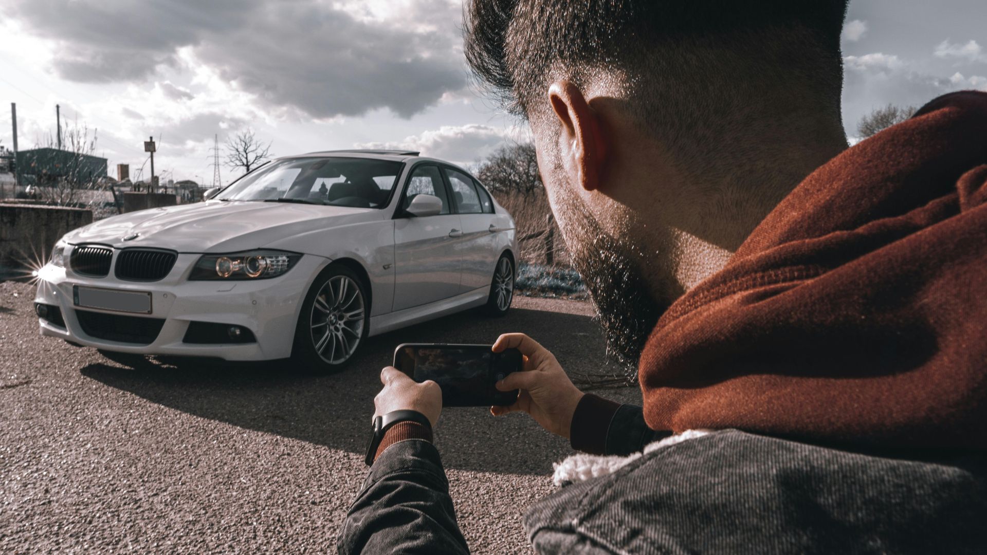 A man takes a picture of a white luxury car using a smartphone outdoors under a cloudy sky.