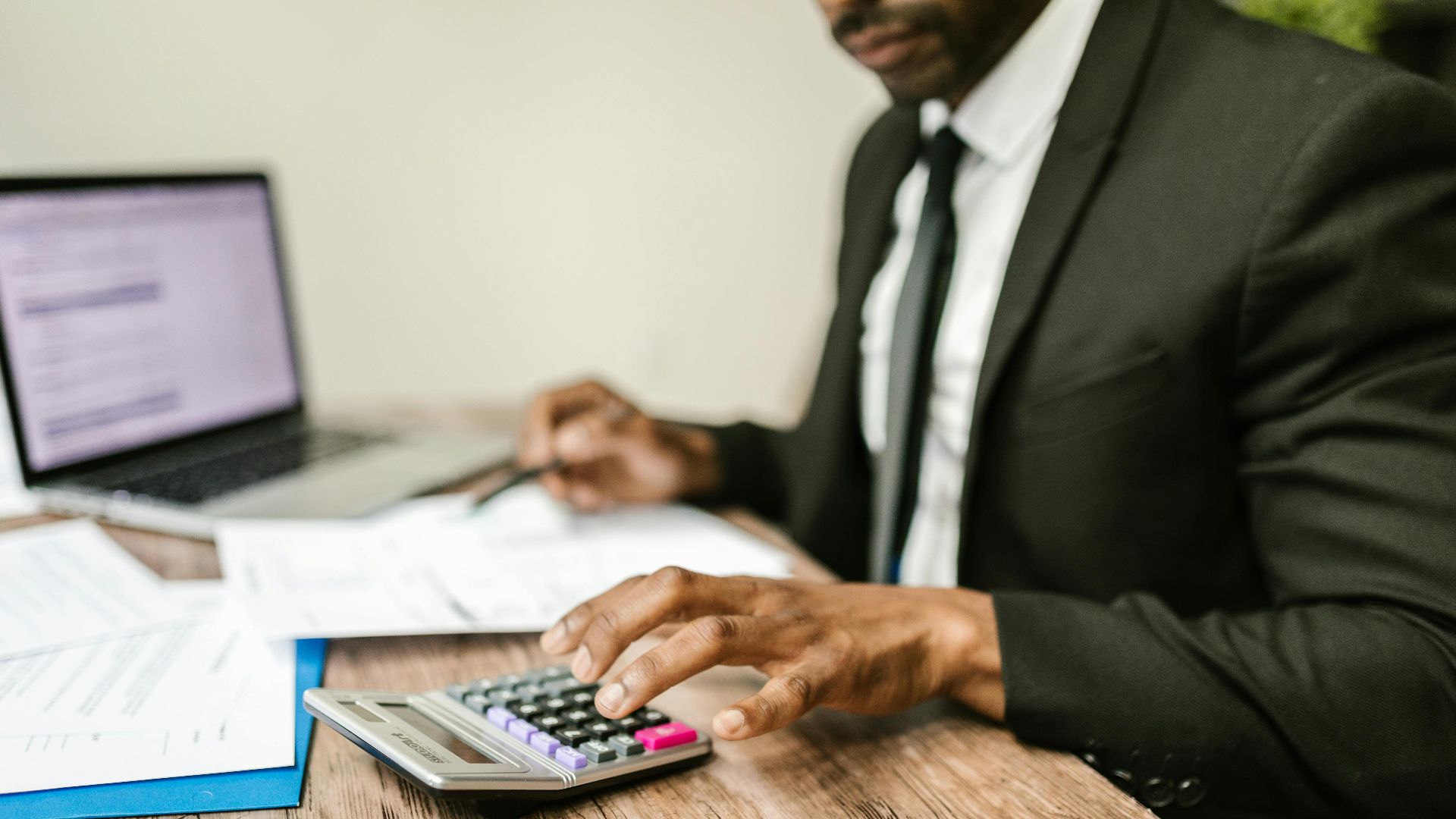 A businessman in a black suit using a calculator at a desk with financial documents and a laptop.