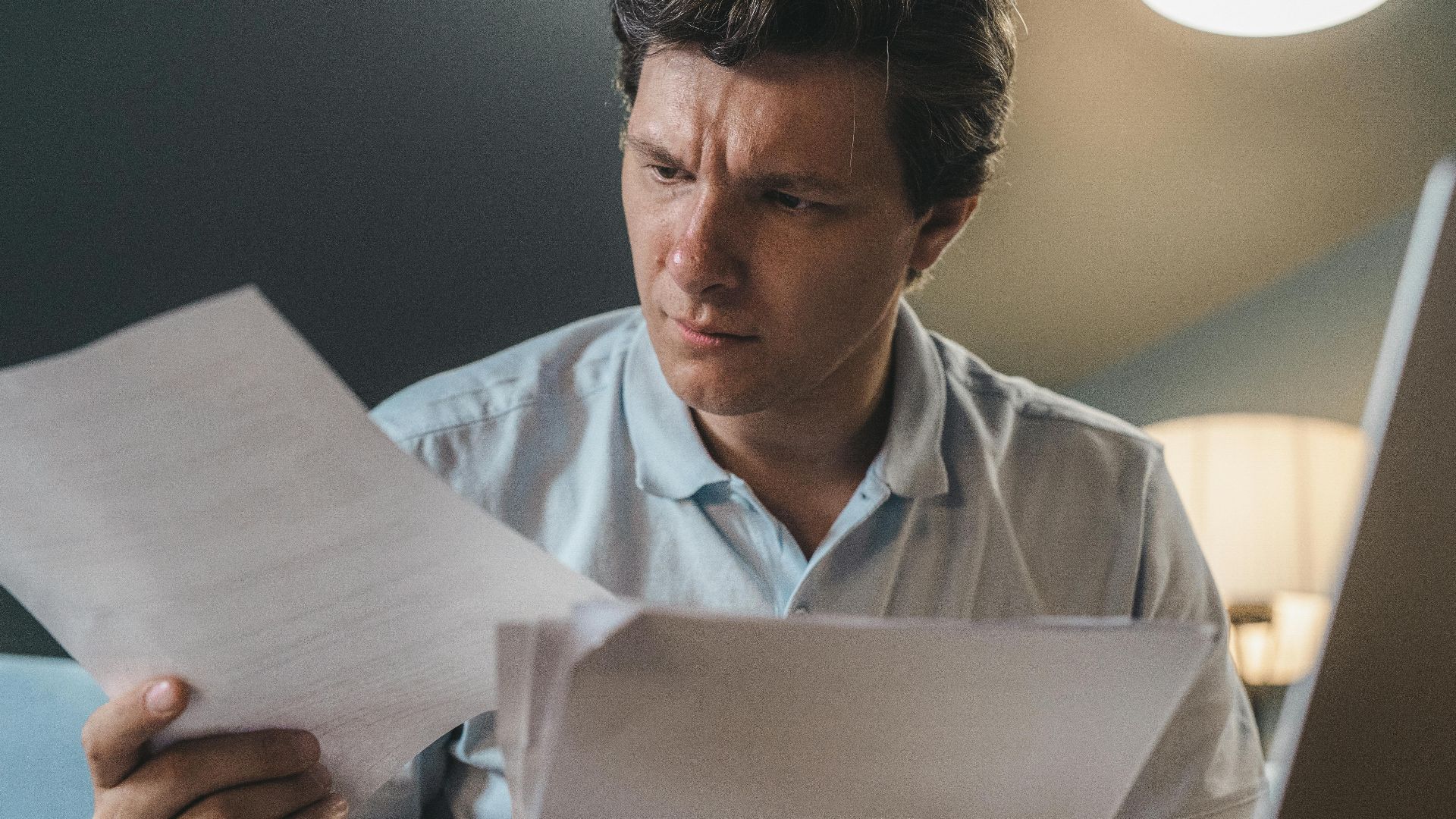 A man intently examines papers, seated indoors under warm lighting, focusing on his work.