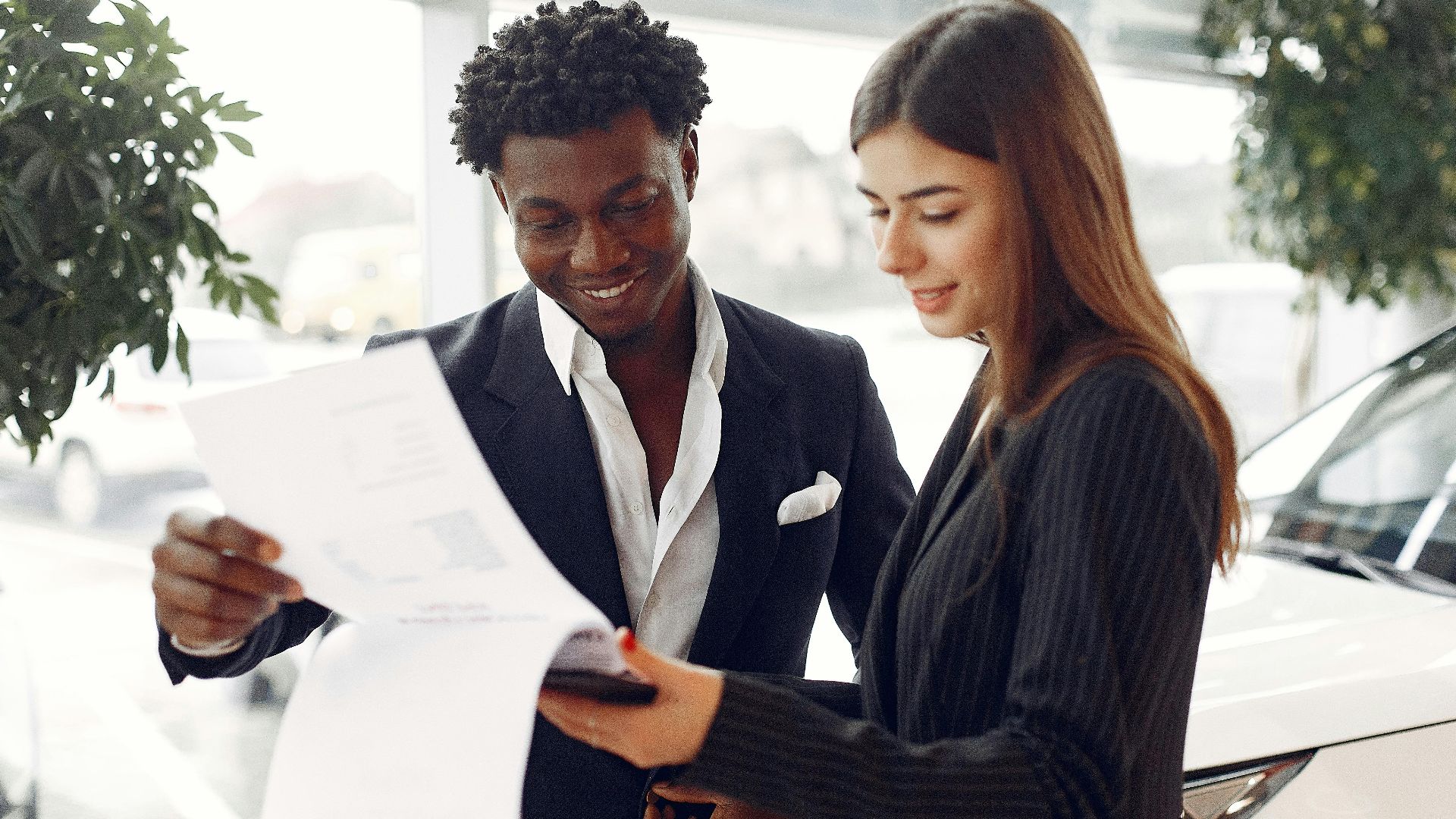 Cheerful smiling multiethnic businessman in classy suit and professional elegant female consultant standing close and reading contract details in car showroom