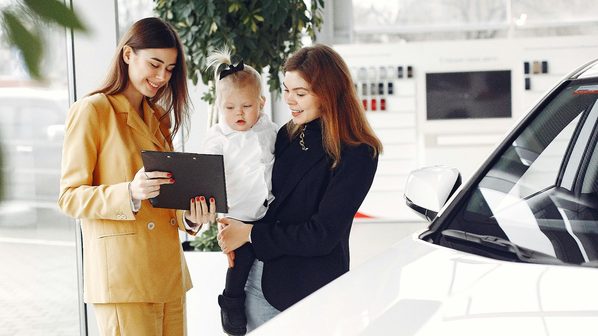 Positive young female with cute little daughter on hands discussing car characteristics with professional dealer in stylish beige suit while standing in car showroom in daylight