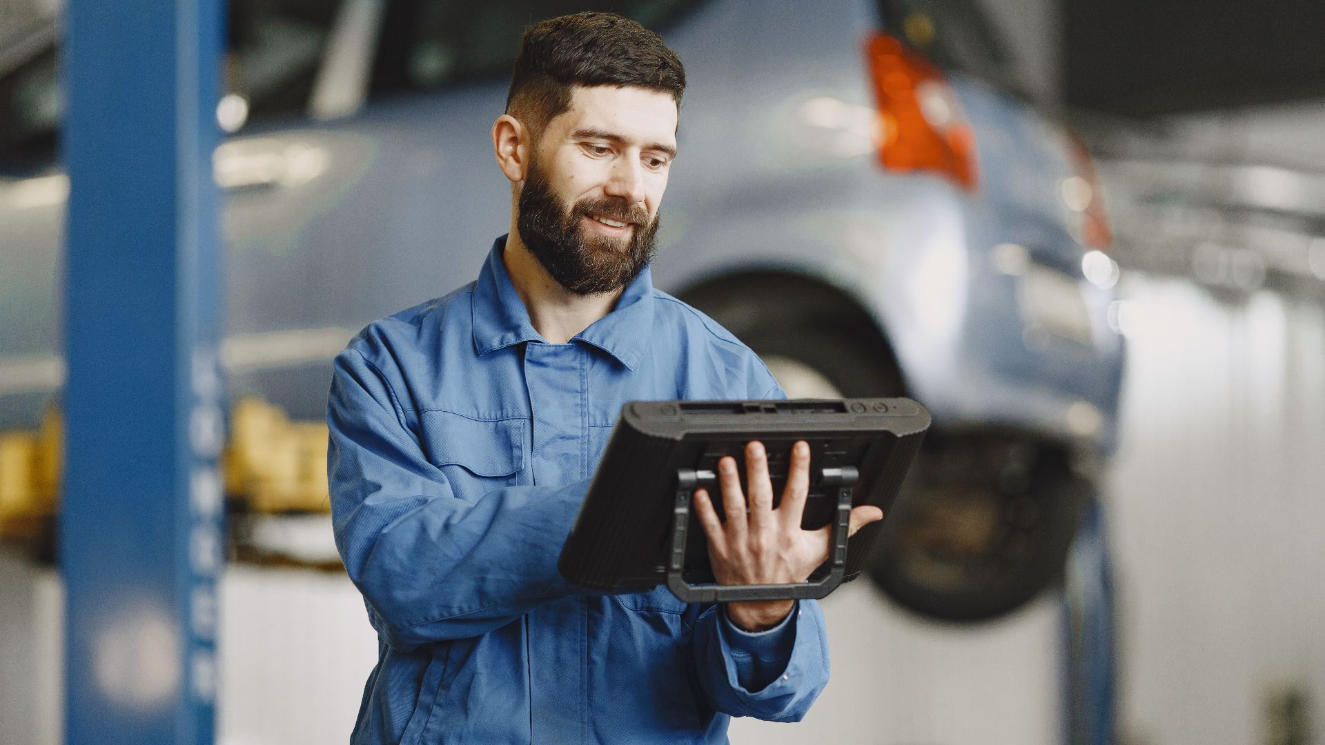 Smiling auto mechanic uses digital tablet in car repair shop environment.
