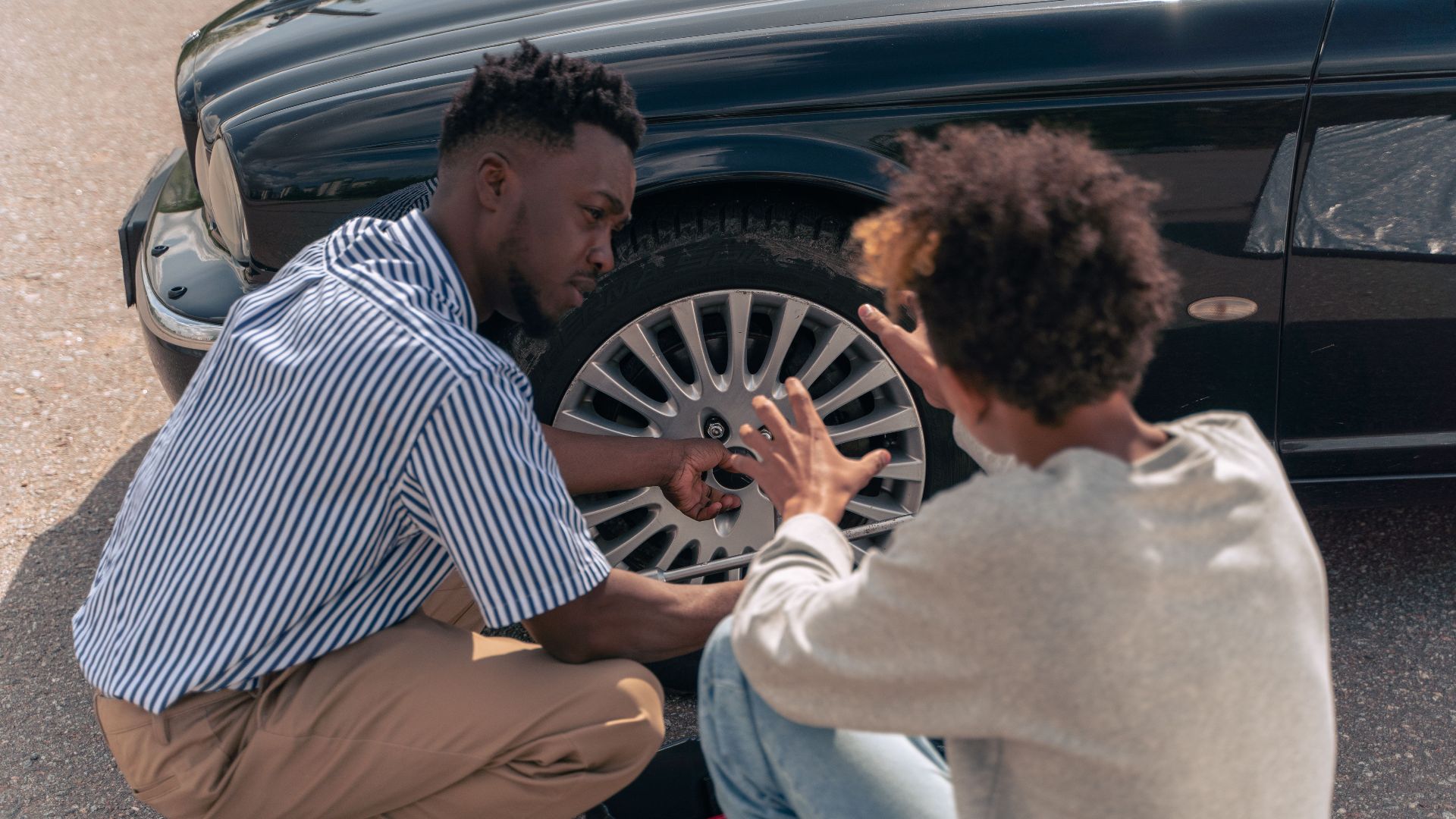A father instructs his teenage son on how to change a car tire outdoors.