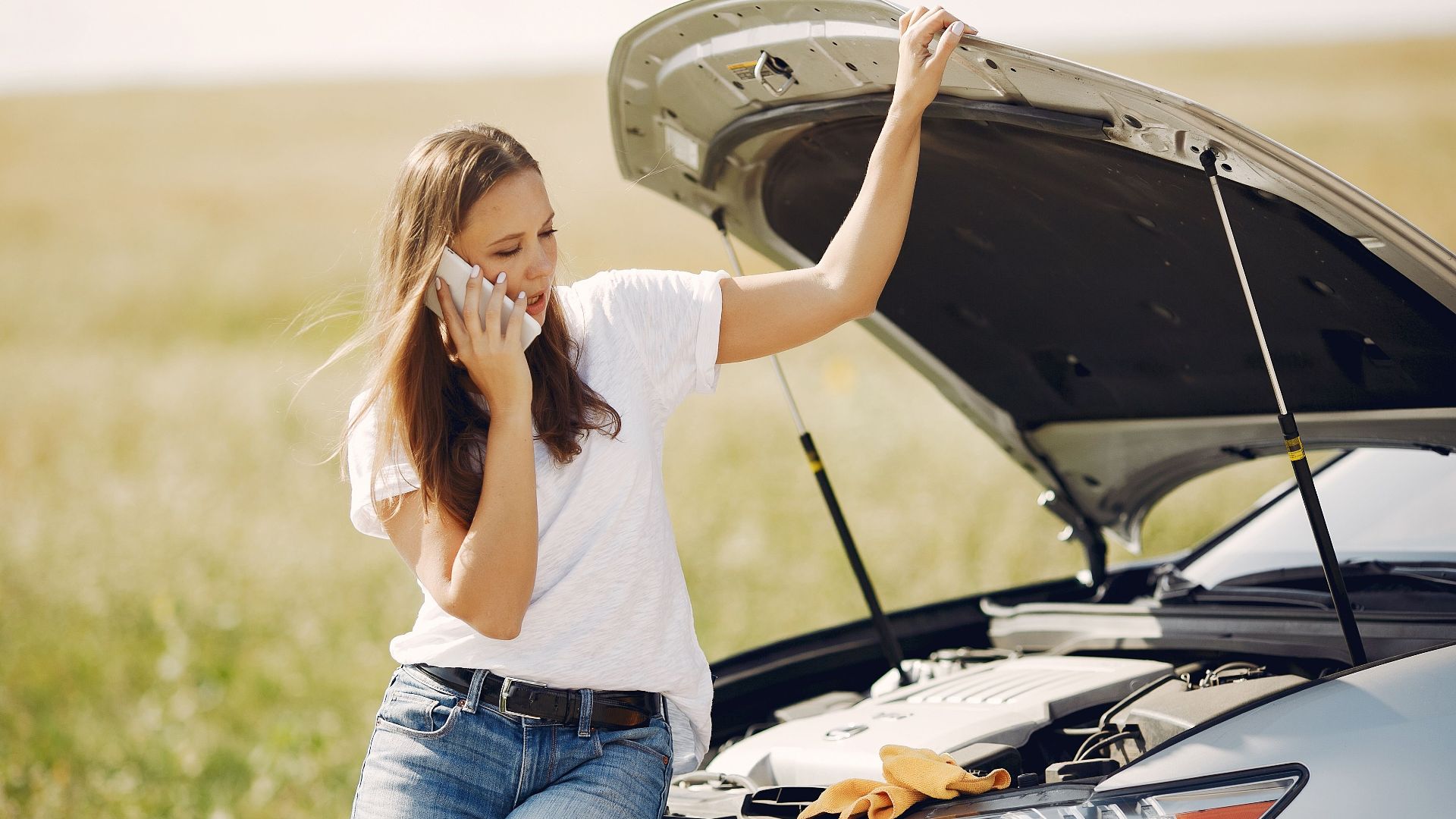 Worried young female driver in white t shirt and jeans speaking on phone while leaning on broken car with open hood asking for help with repair against green field in sunny summer day