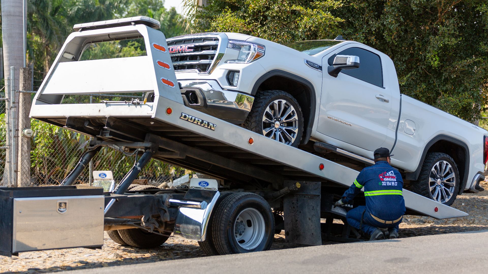 Tow truck operator loading white GMC pickup truck on street in daytime.