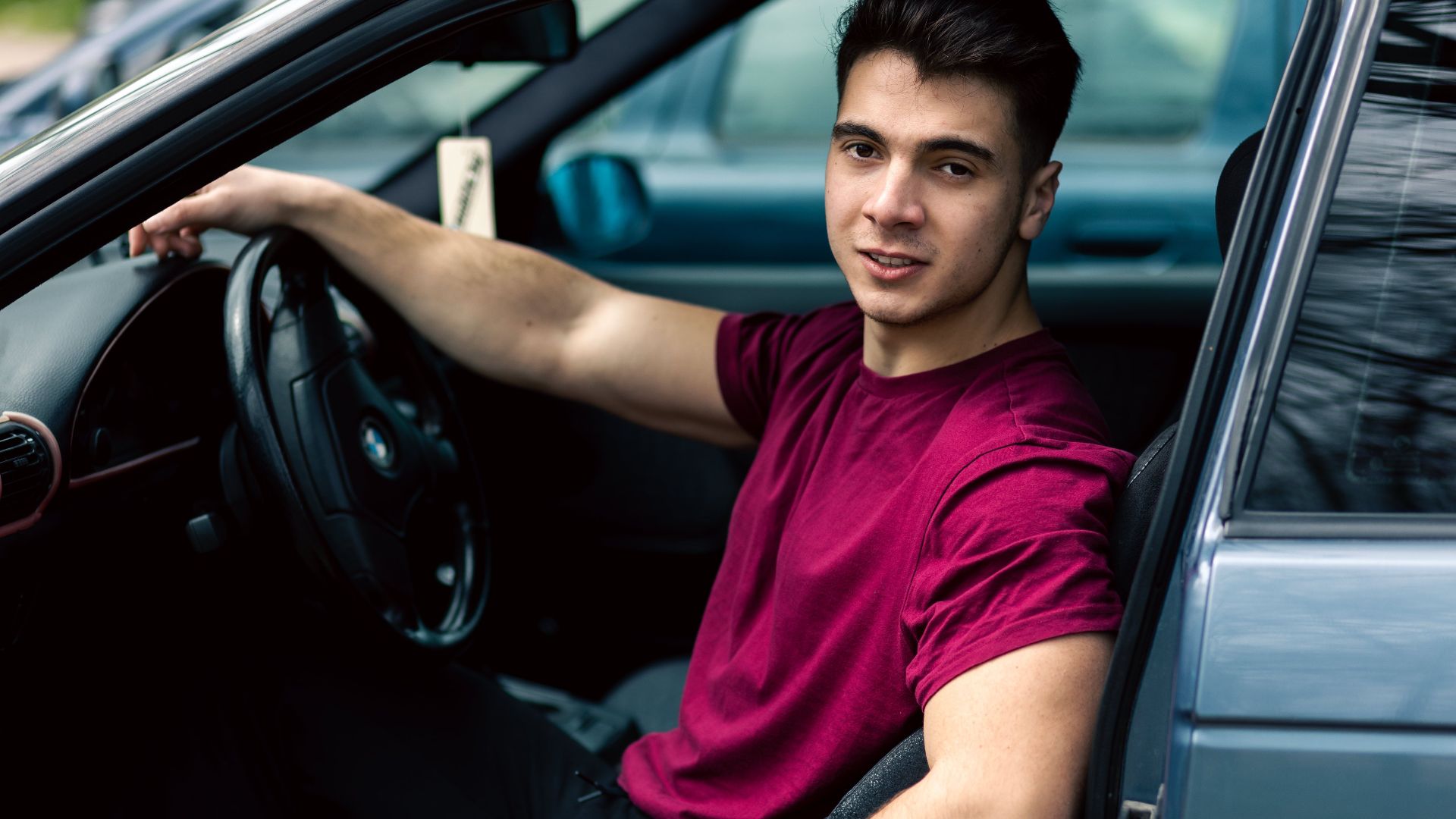 A young man wearing a maroon t-shirt sits casually in a blue car, portraying a relaxed and modern lifestyle.
