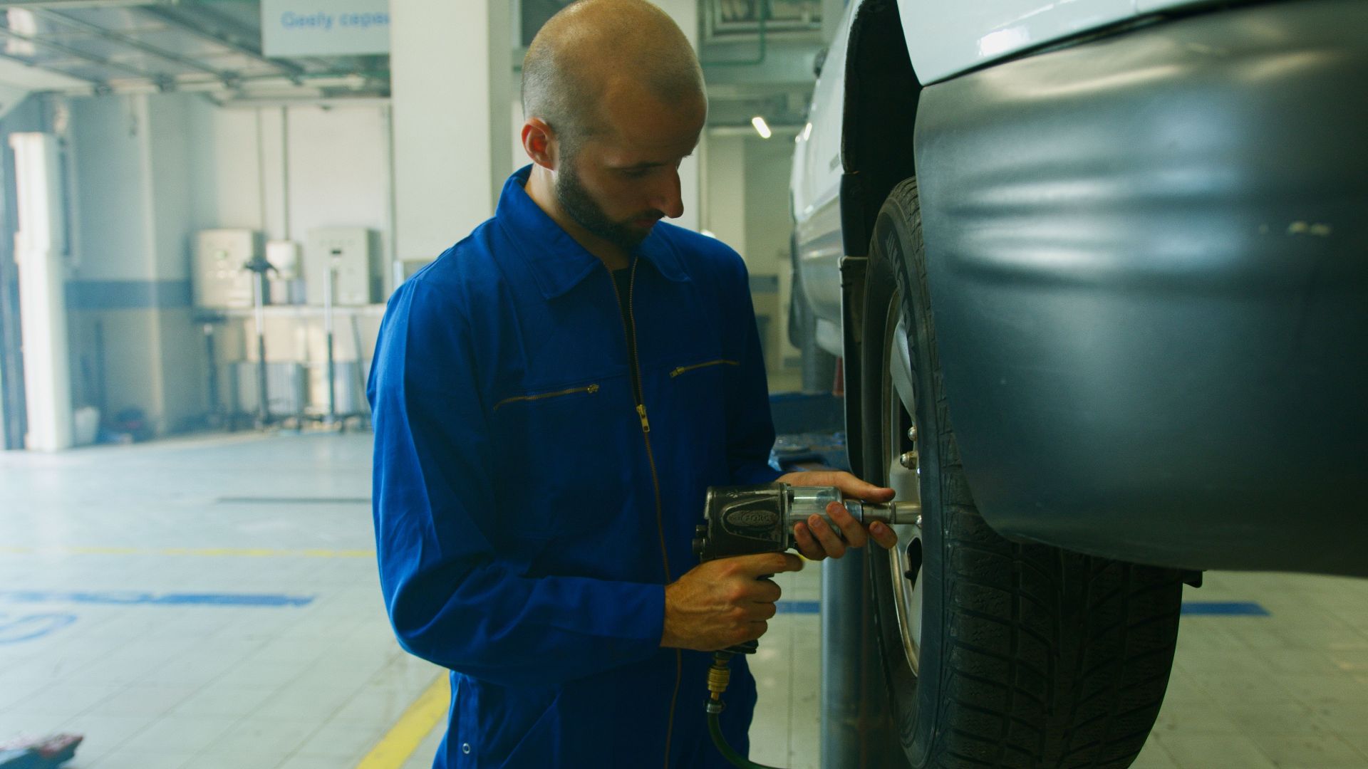 Mechanic in blue uniform servicing a car with an impact wrench in a garage.