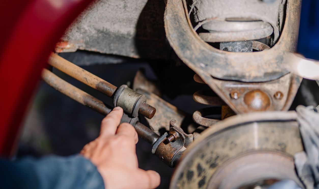 Mechanic’s hand pointing at a worn car suspension bushing and rusted linkage near the wheel assembly during a vehicle inspection or repair process.
