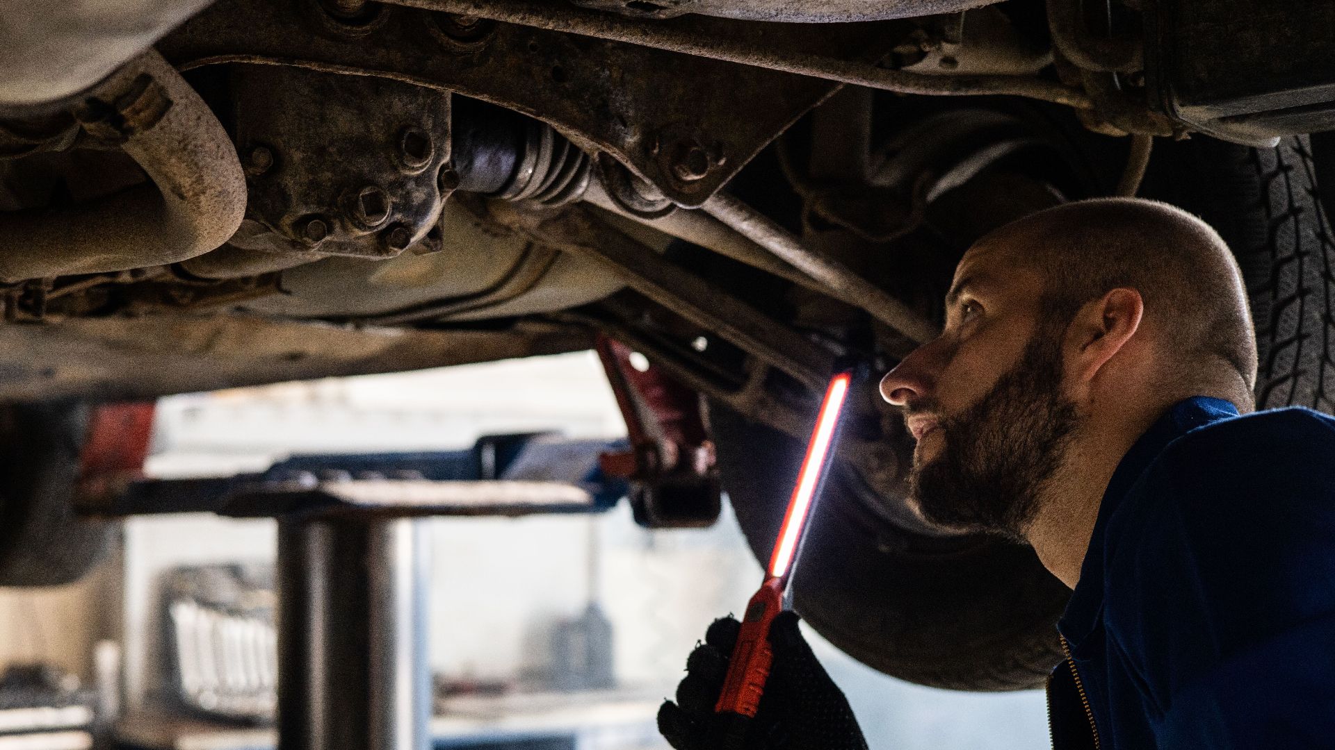 Mechanic using light to inspect vehicle undercarriage in workshop for maintenance.