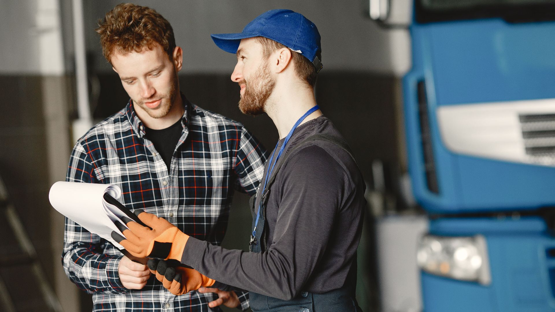 Mechanic in uniform consults with customer in garage setting beside a blue truck.