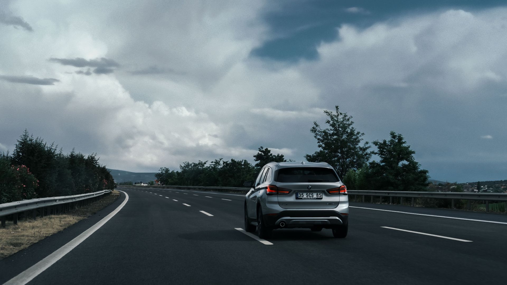 A silver SUV travels on a highway surrounded by lush greenery and dramatic clouds.