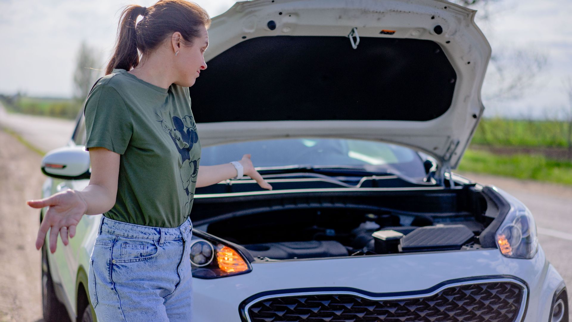 A woman looks puzzled by her broken car with an open hood on a rural road.