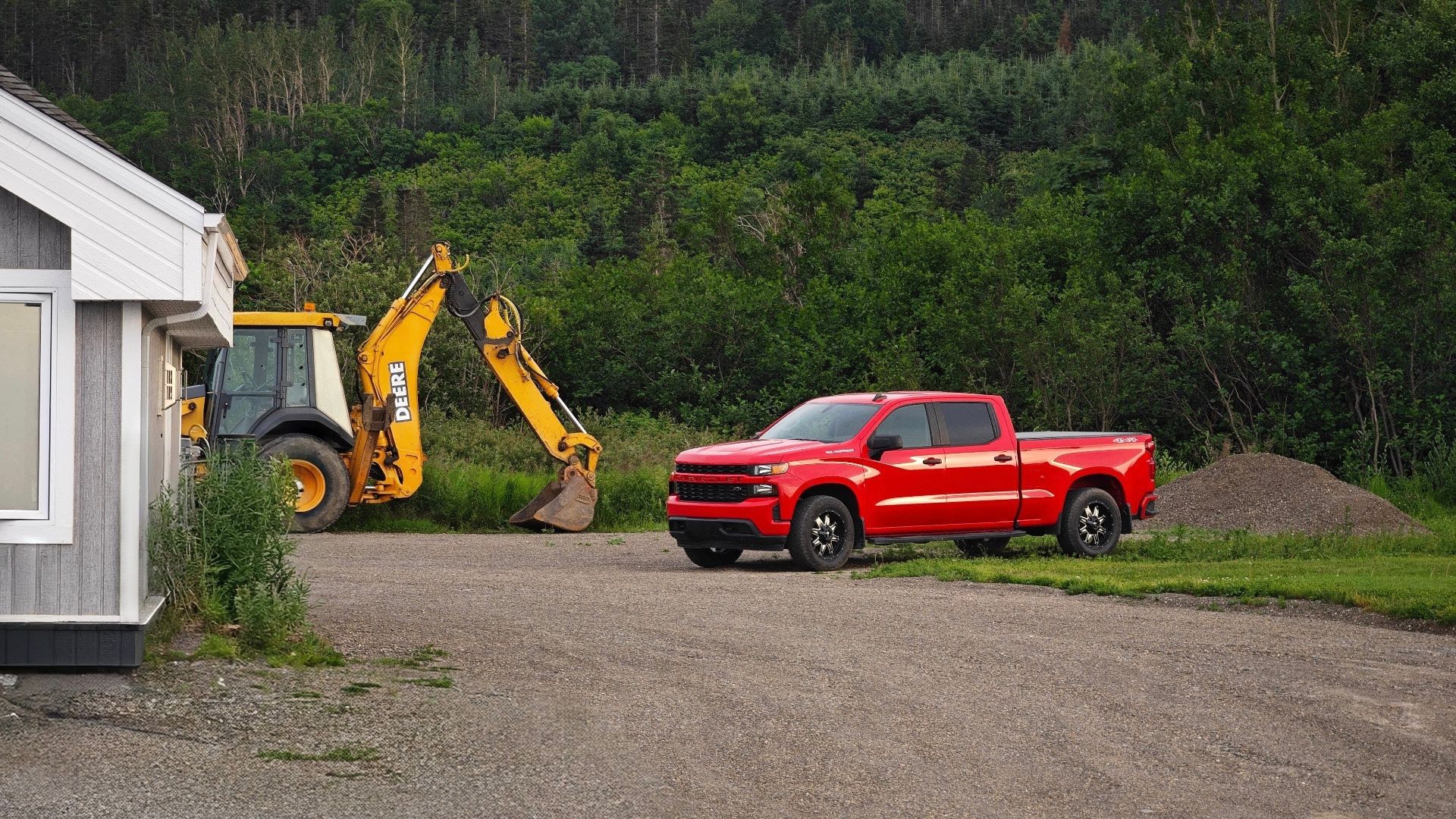 A red pickup truck and yellow backhoe parked in a rural driveway with lush forest backdrop.