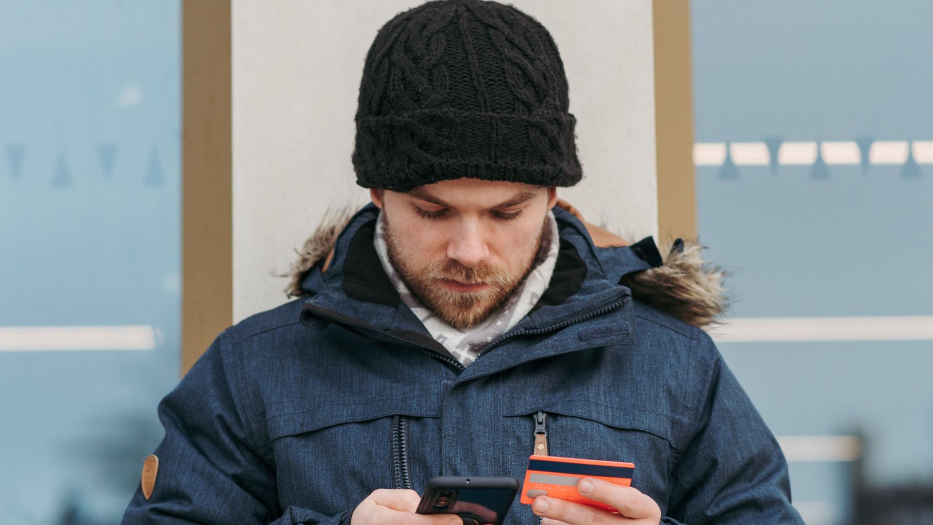 Serious young guy in warm clothes and hat standing on city street and browsing smartphone while making online payment with credit card near modern building