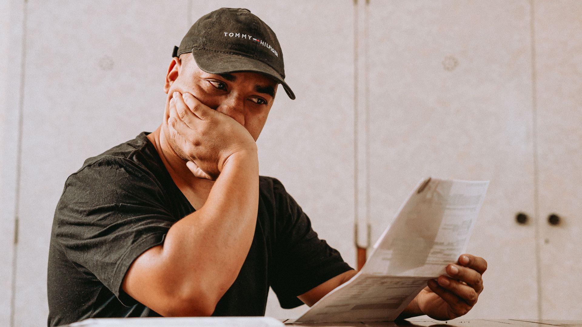 A man sits indoors, deeply focused on reviewing paperwork, expressing concern and contemplation.