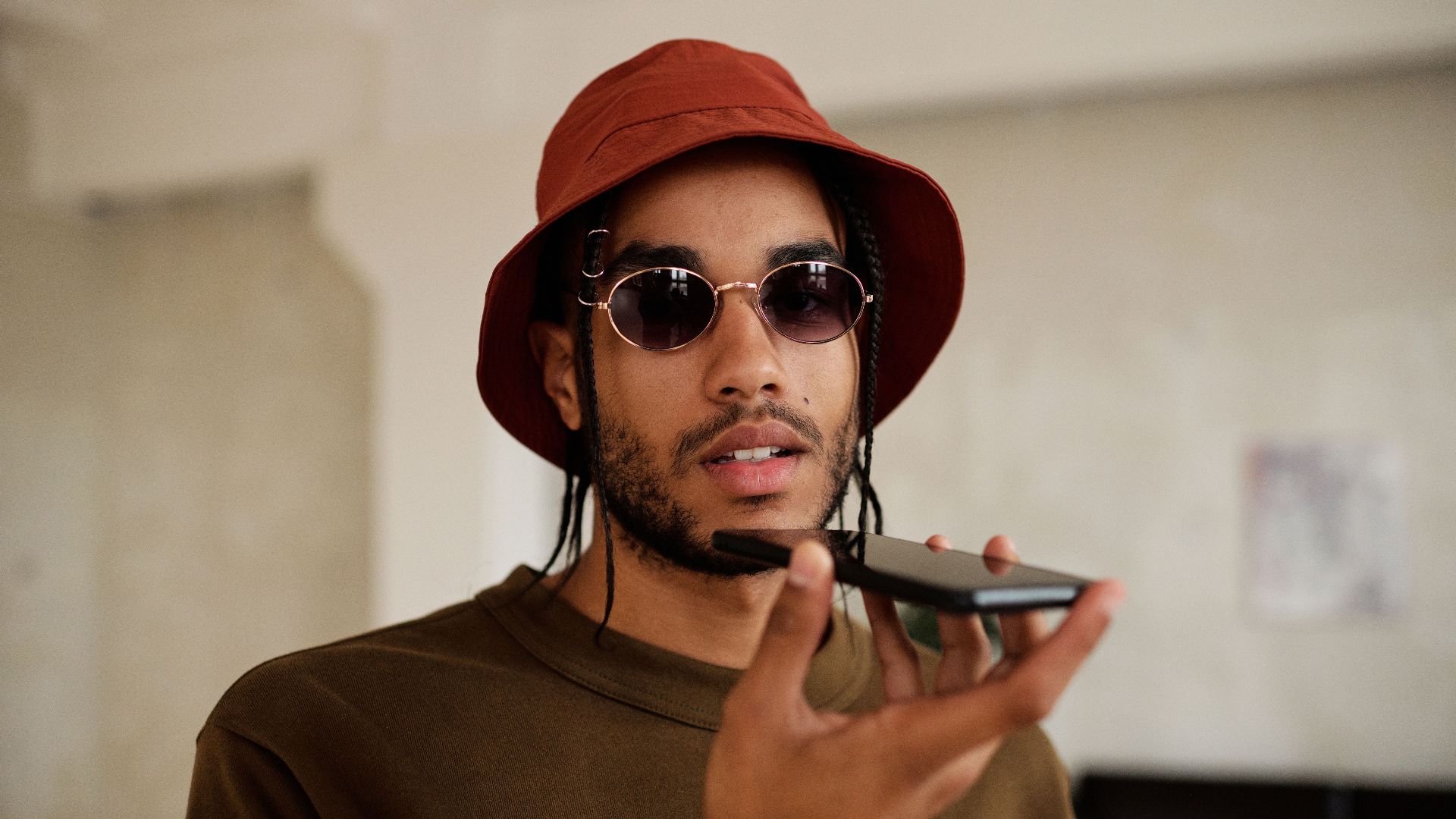 Fashionable man wearing sunglasses and a bucket hat speaking on a smartphone indoors.