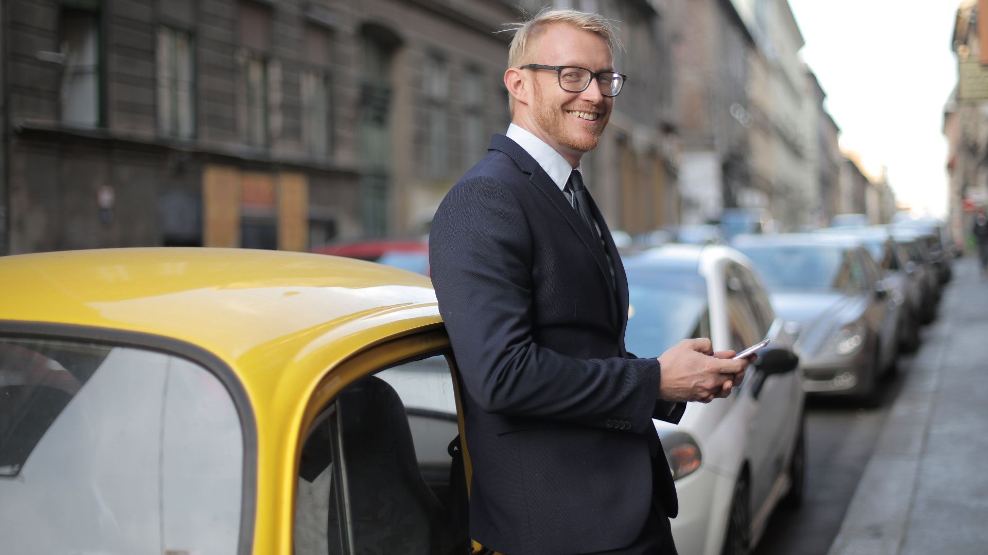A stylish businessman in formal attire stands confidently by a yellow car on a bustling city street.