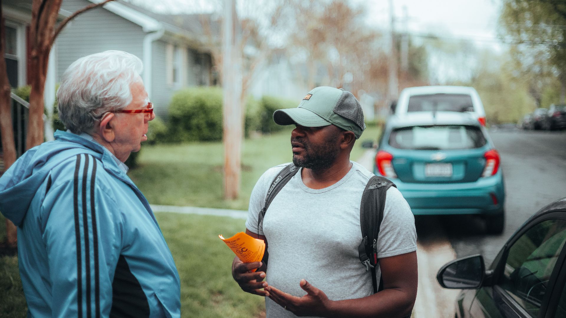 Two men engaged in conversation on a suburban sidewalk with parked cars.