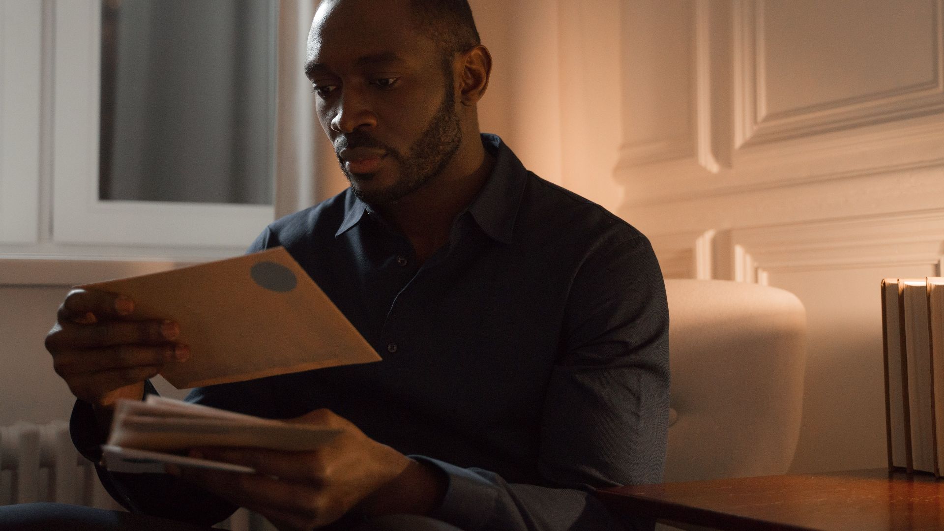 A man with a mustache reads letters while sitting in a warmly lit home interior.