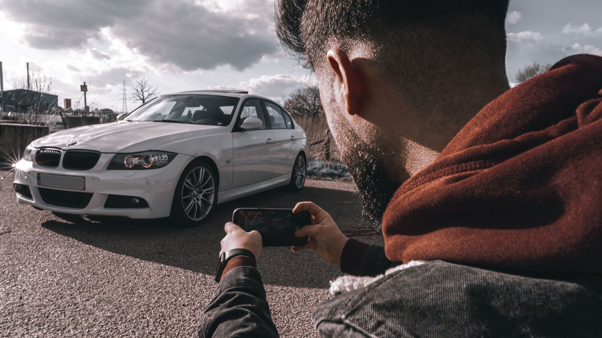 A man takes a picture of a white luxury car using a smartphone outdoors under a cloudy sky.