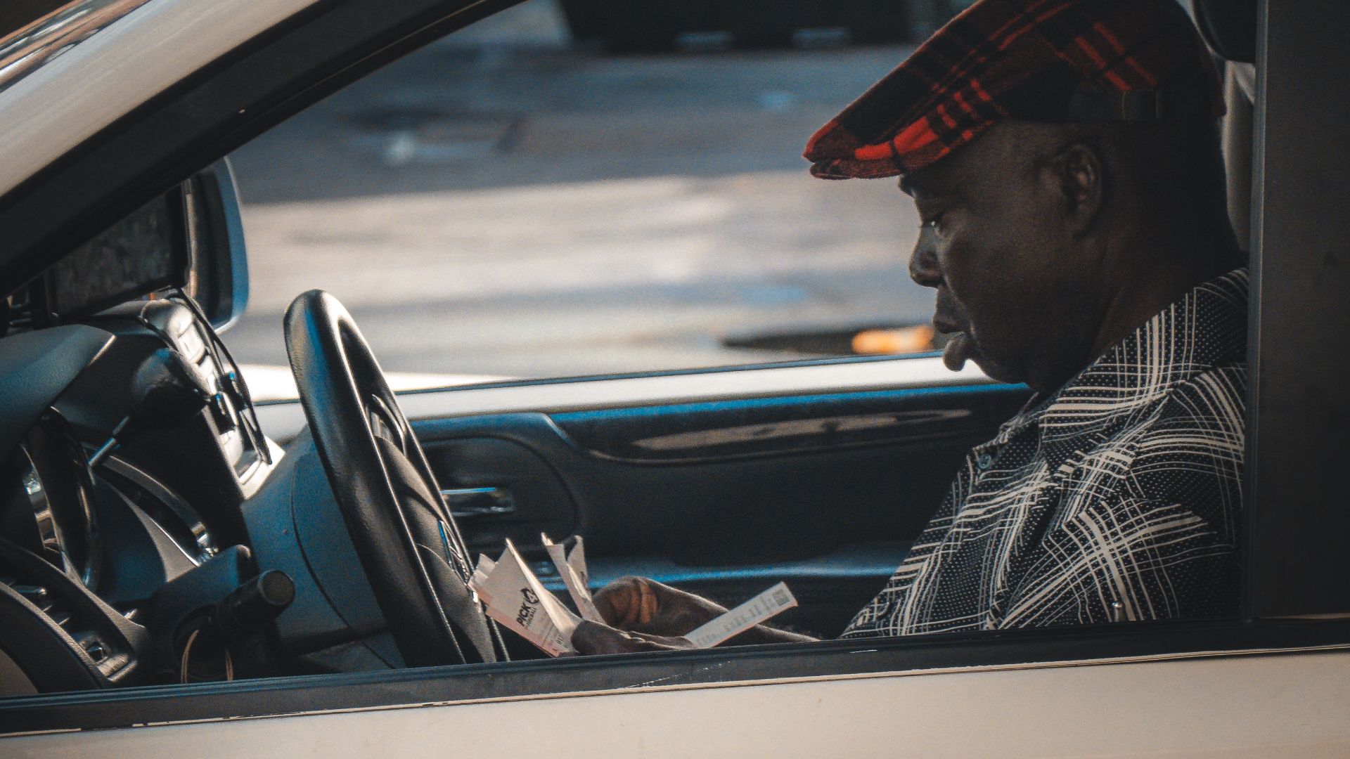 An adult man reads a newspaper inside a parked car during daytime.
