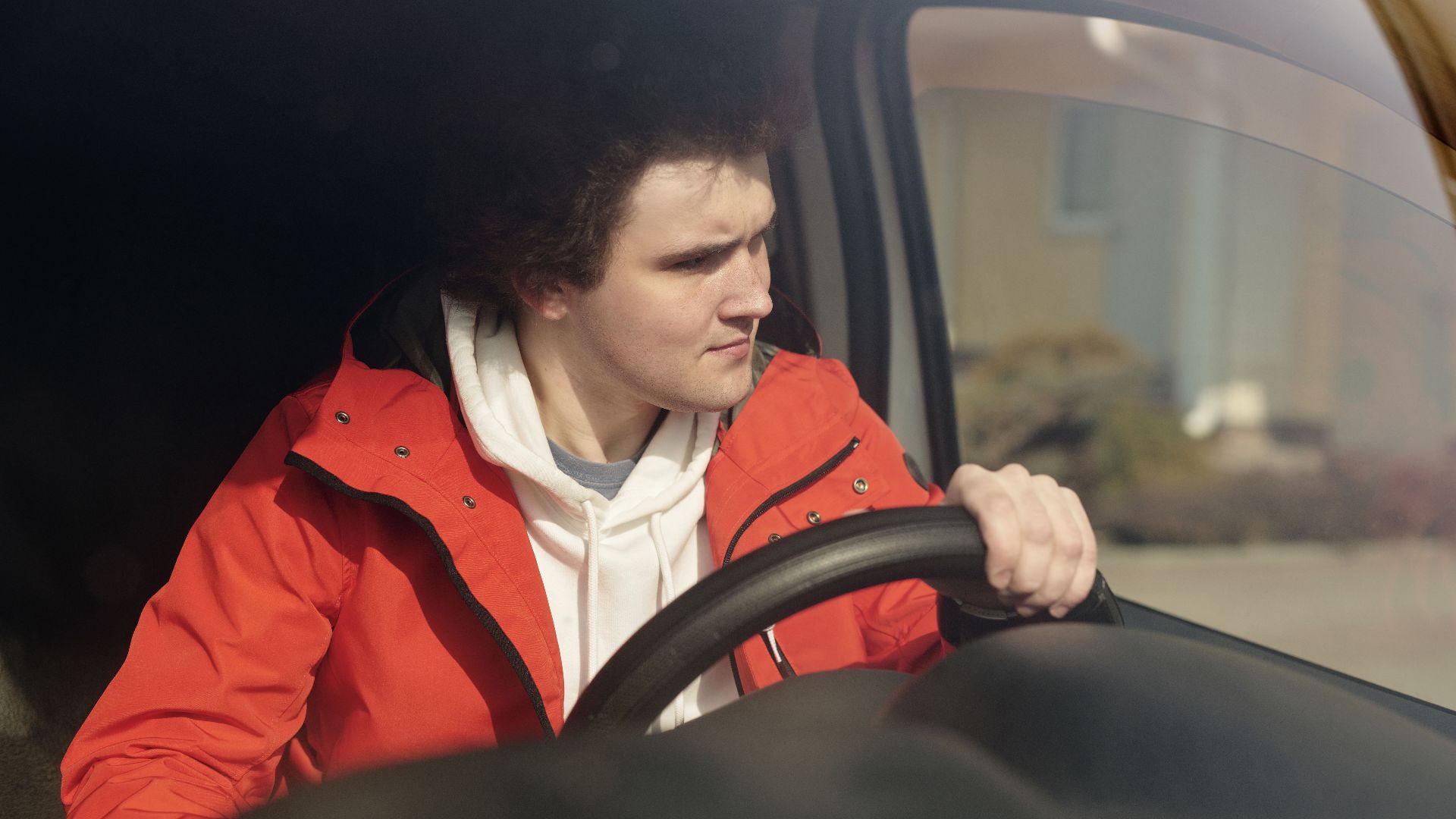 A young man in a red jacket steering a car with focus, enjoying a peaceful drive.