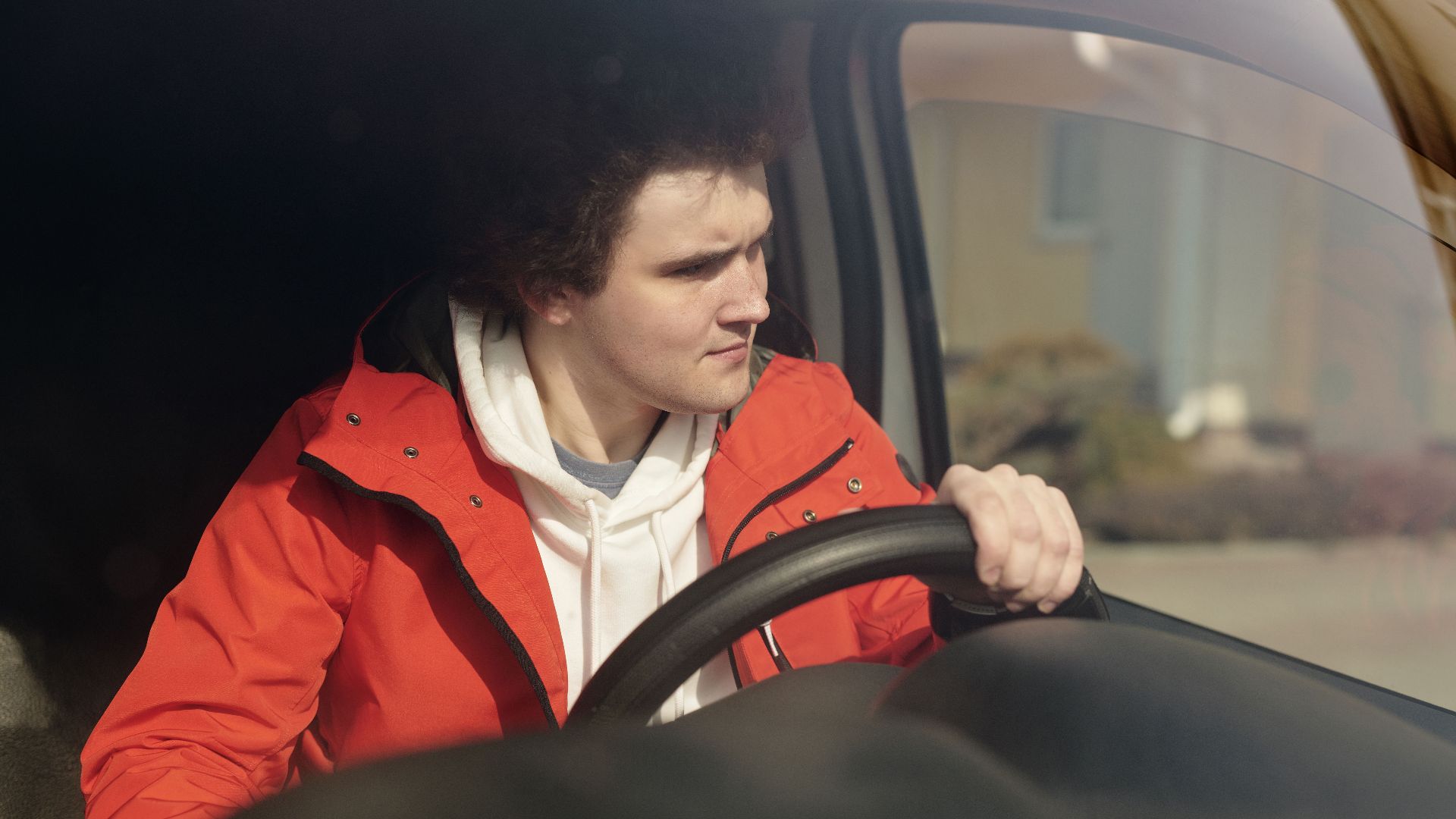 A young man in a red jacket steering a car with focus, enjoying a peaceful drive.