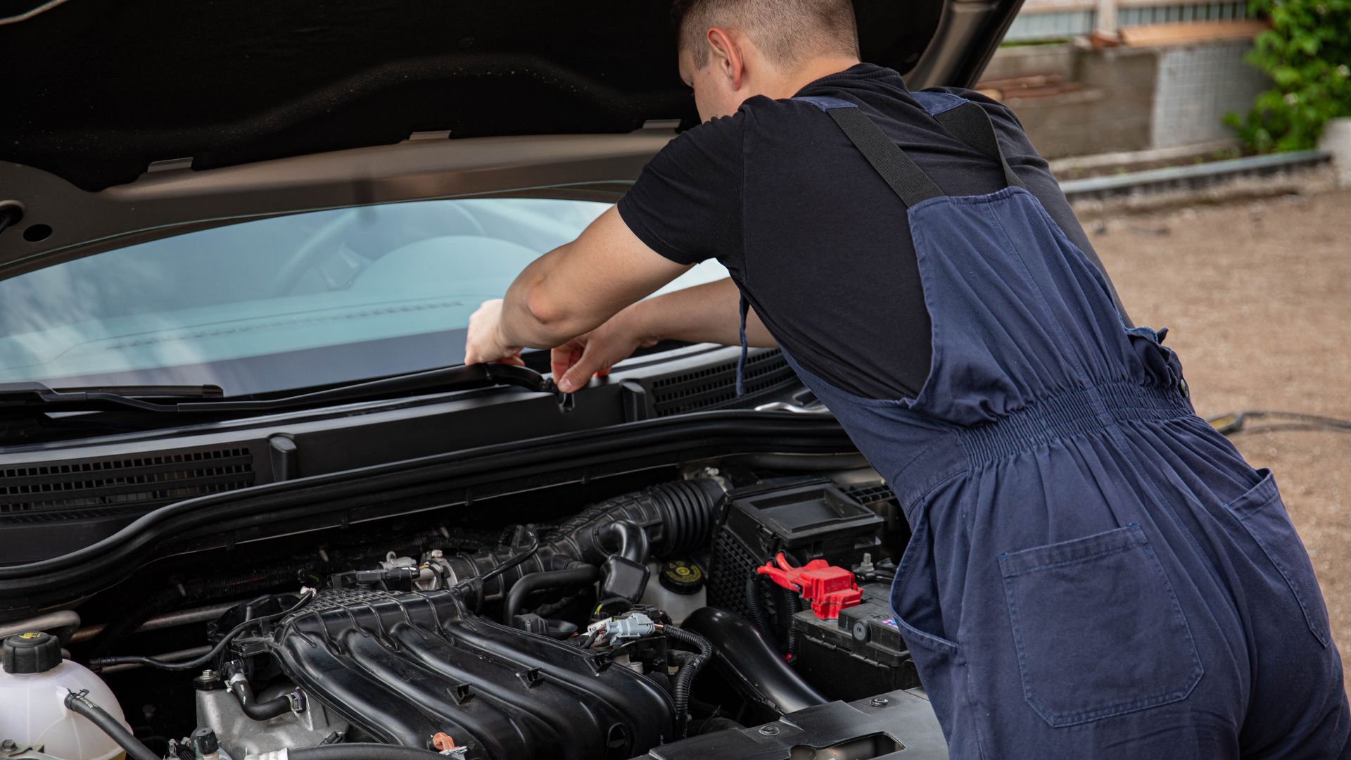 A mechanic in blue overalls works on a car engine outdoors, showcasing vehicle maintenance skills.