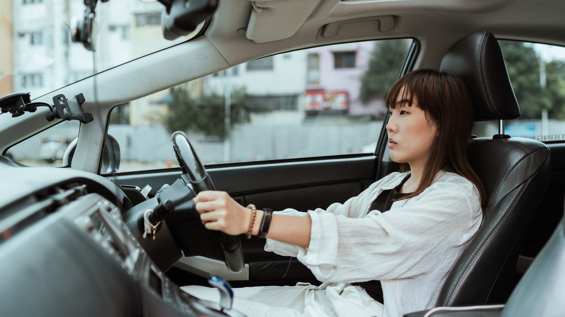 Side view unemotional Asian female in casual white clothes steering right hand drive car along urban streets in daytime