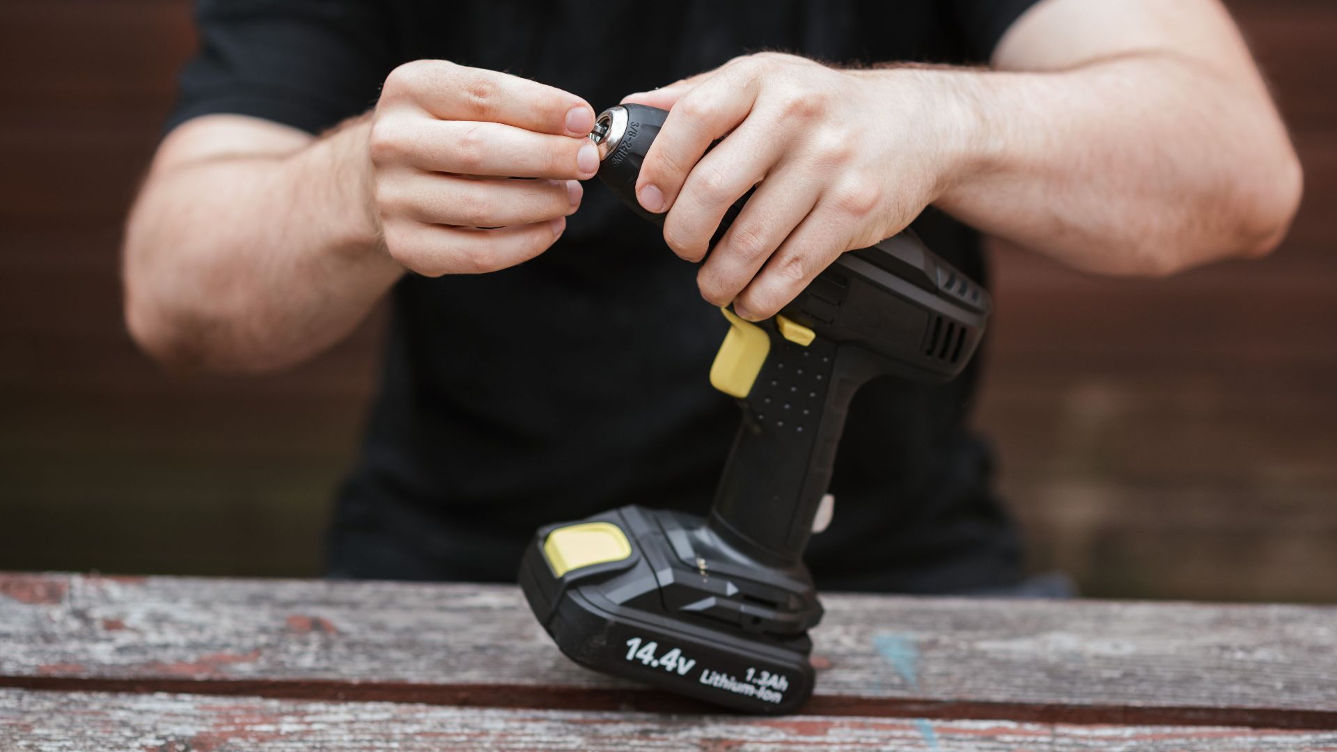 Crop anonymous skilled master in casual outfit preparing electric drill for job on wooden table in soft daylight