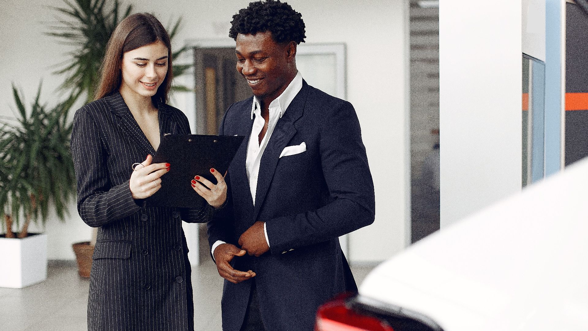 Cheerful multiethnic stylish female agent and smiling customer in formal suit discussing contract details while standing in modern car showroom in daylight