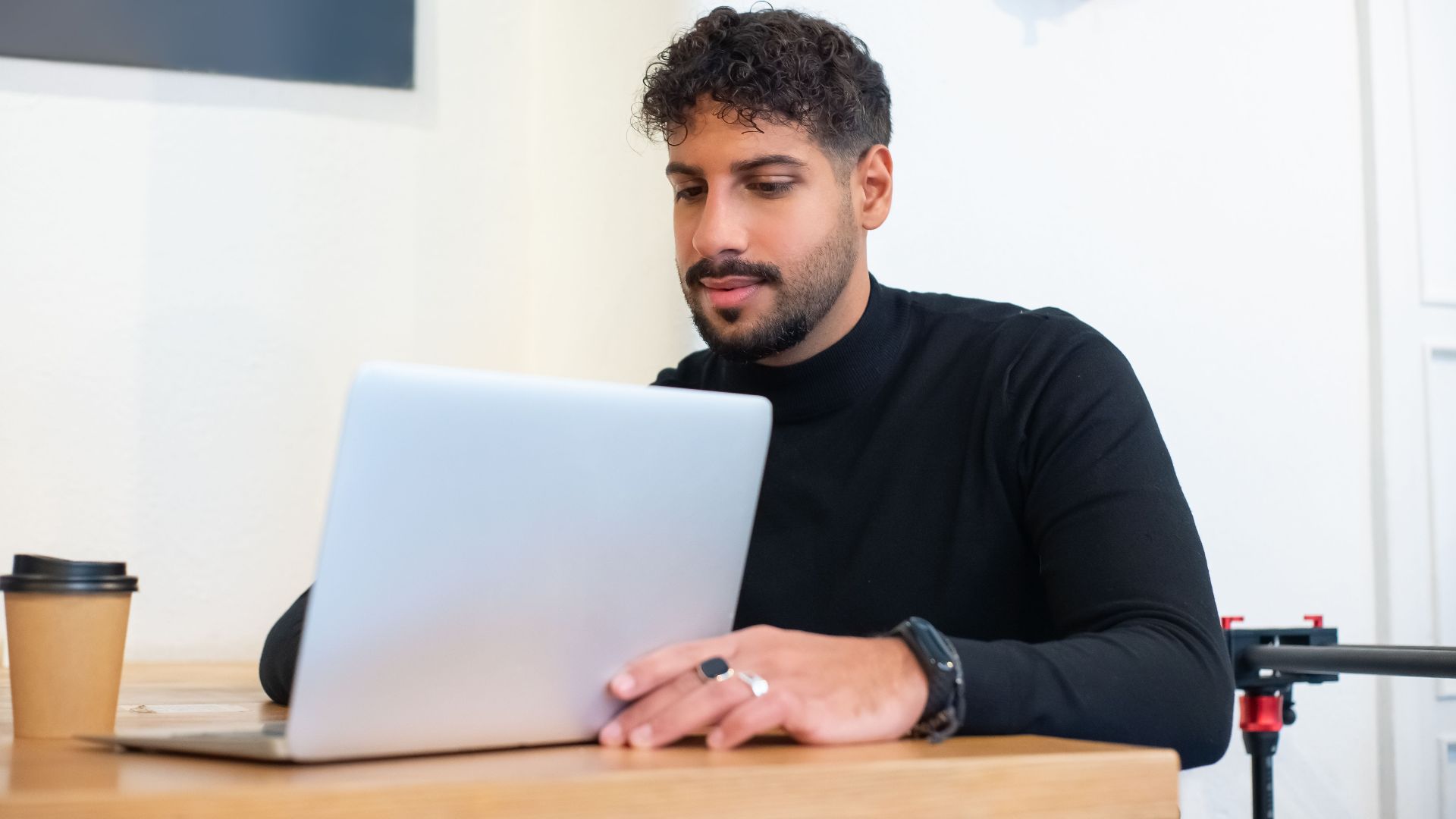 Bearded man in a black sweater using a laptop in a contemporary office setting.