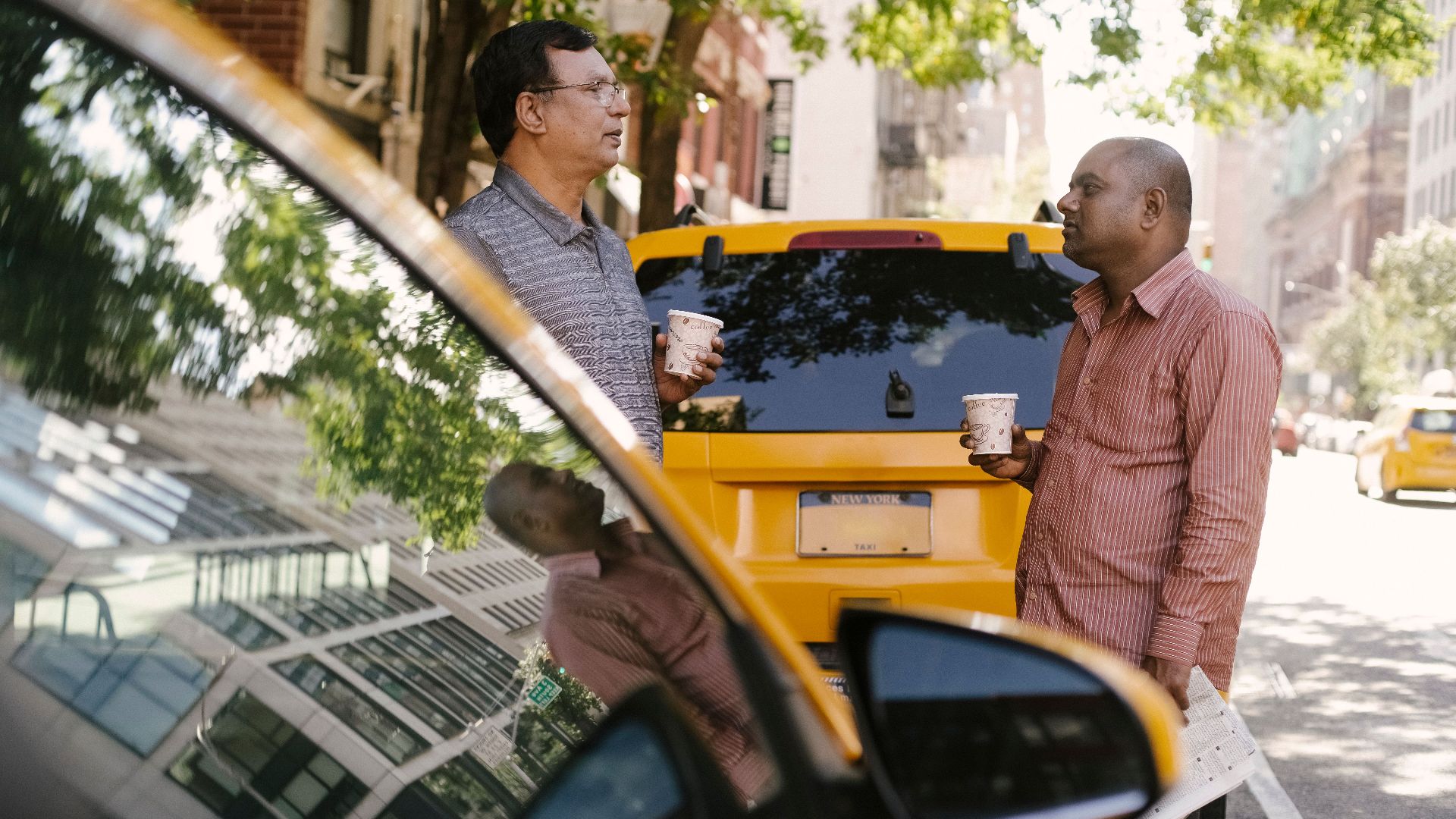 Side view of multiracial male coworkers with takeaway coffee conversing near yellow automobiles on urban road while looking at each other