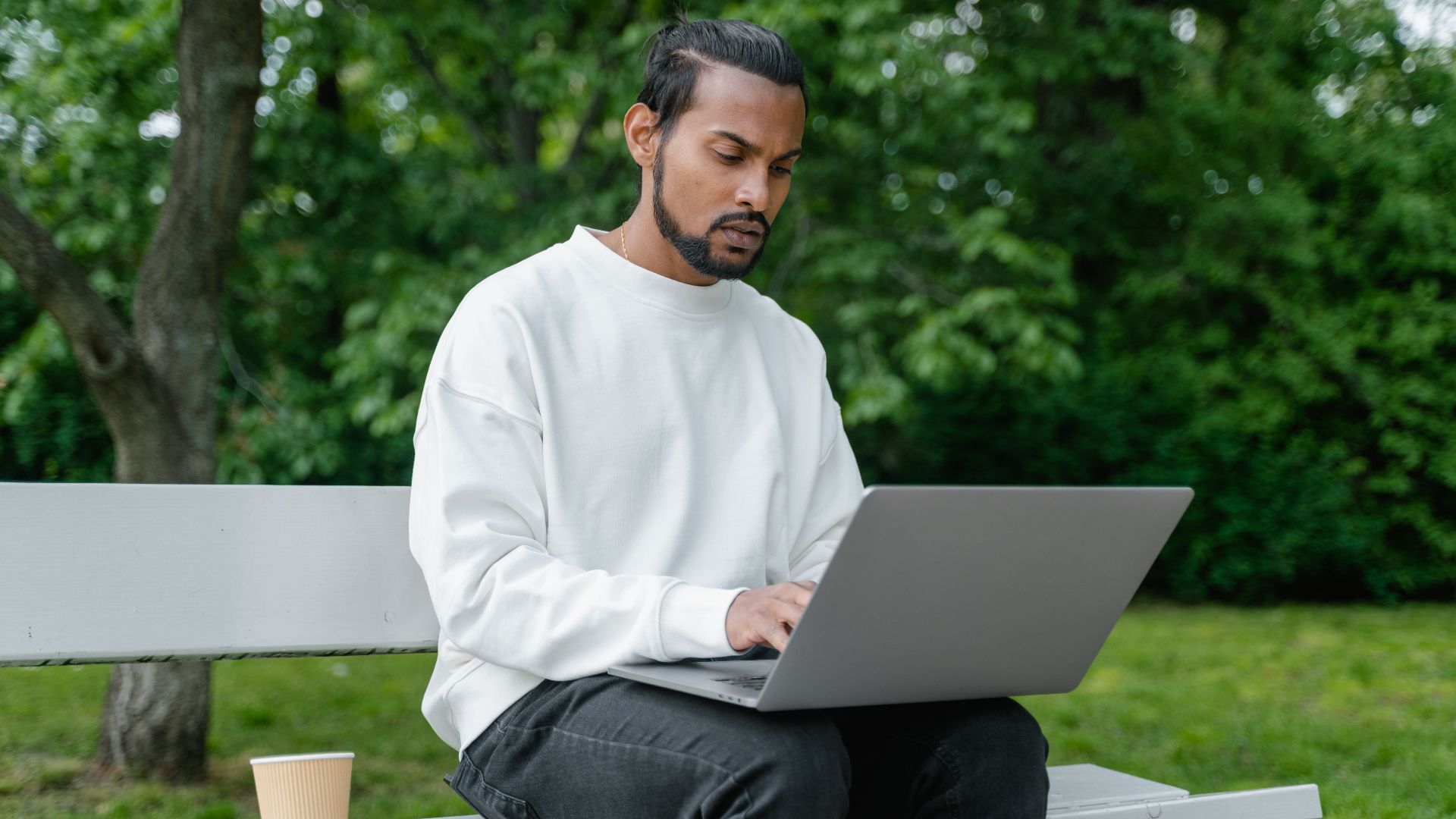 Man in white sweater working on a laptop outdoors on a park bench with greenery around.