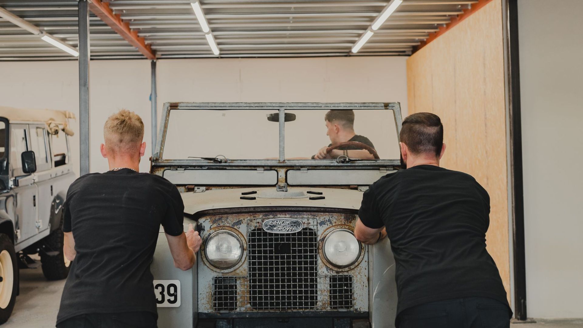 Two men push a classic Land Rover Series vehicle into a modern garage, showcasing teamwork and vintage style.
