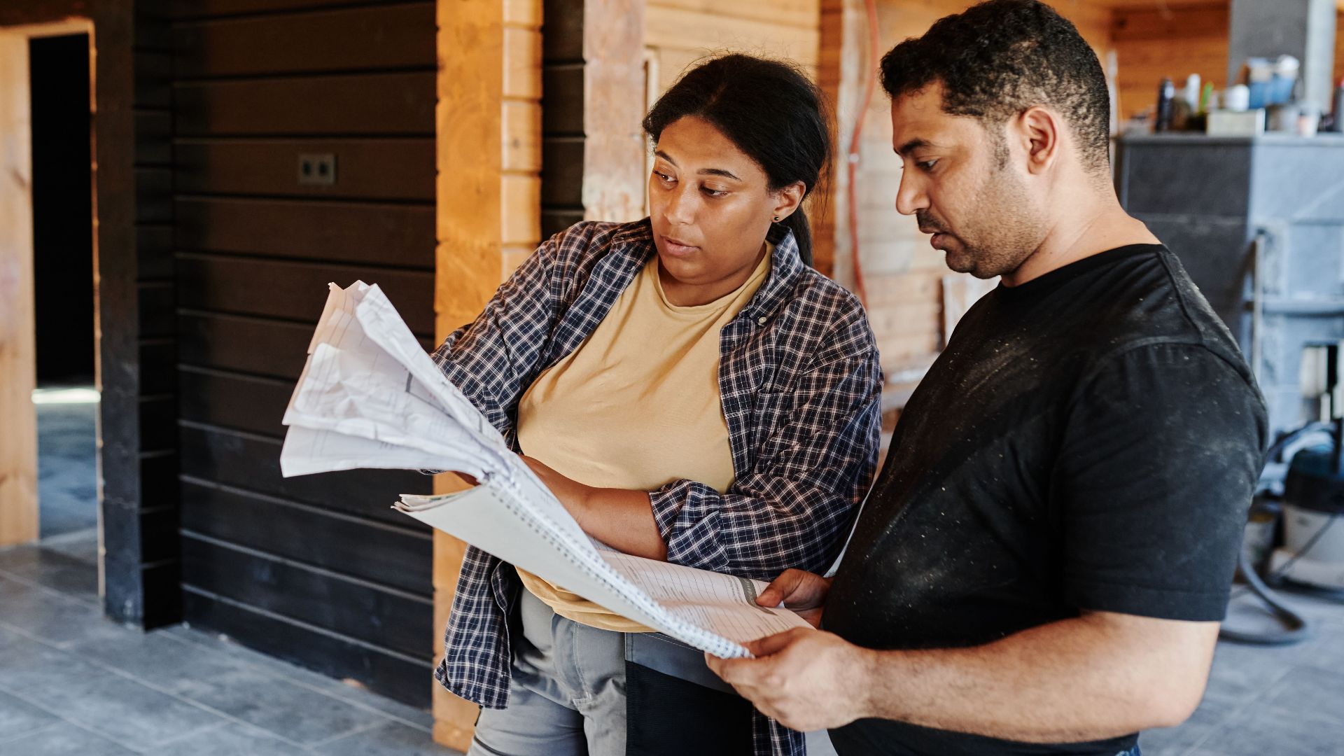 Two construction workers review blueprints inside a wooden structure, discussing plans.