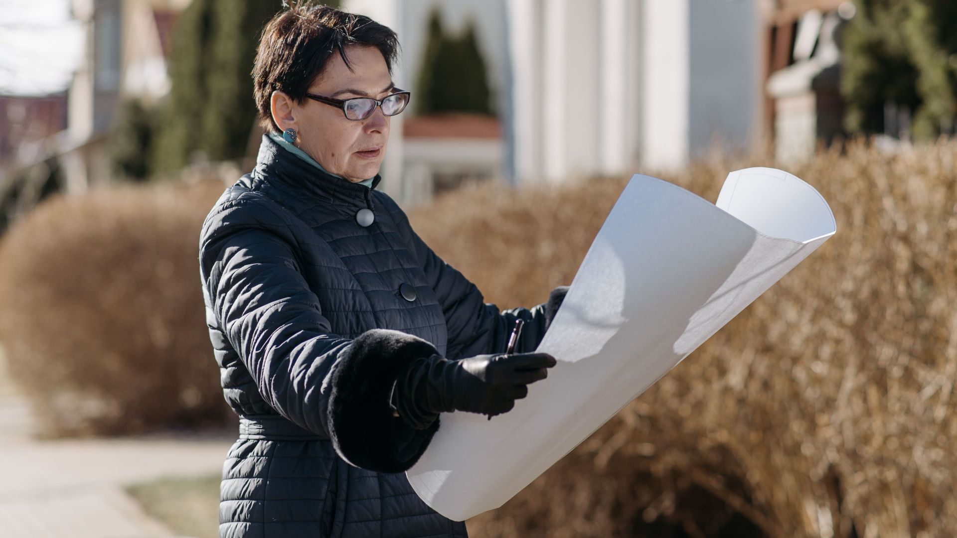 Woman examining a blueprint in a sunny residential area, considering properties.