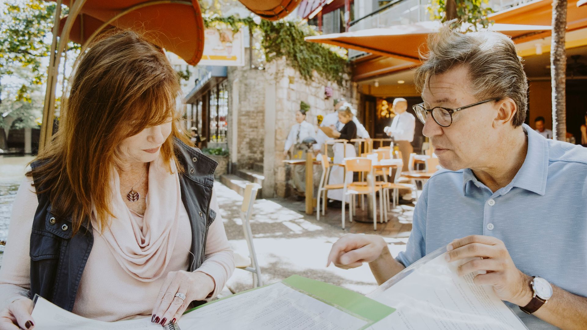 man and woman sitting down near table while reading outside cafe during daytime