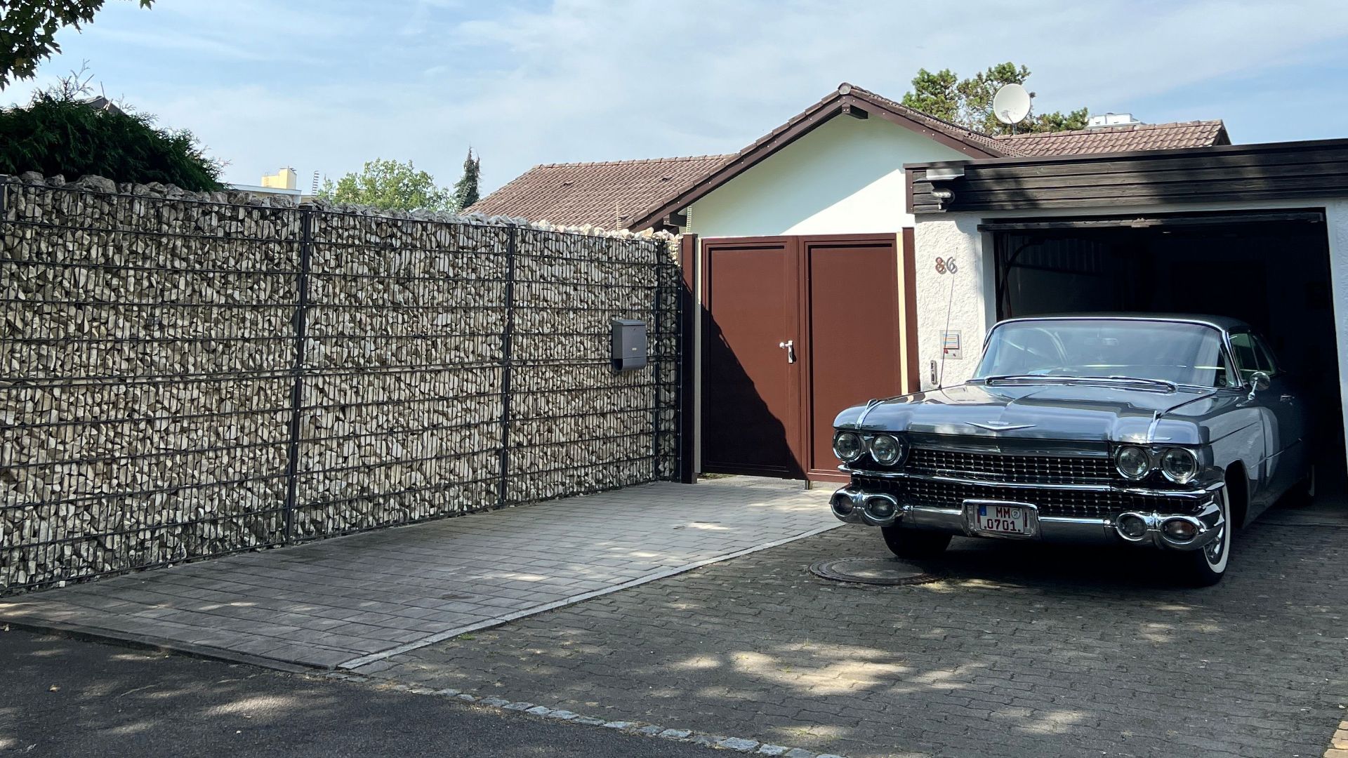 A classic vintage car parked beside a stone wall and garage under a clear blue sky.