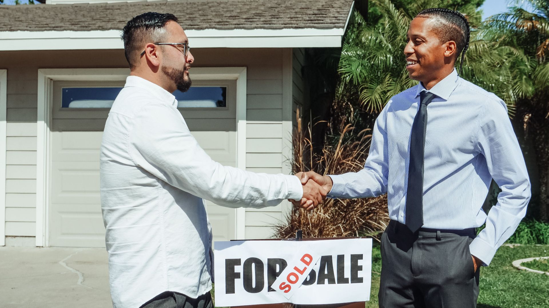 Two men shaking hands in front of house sold sign, sealing real estate deal outdoors.