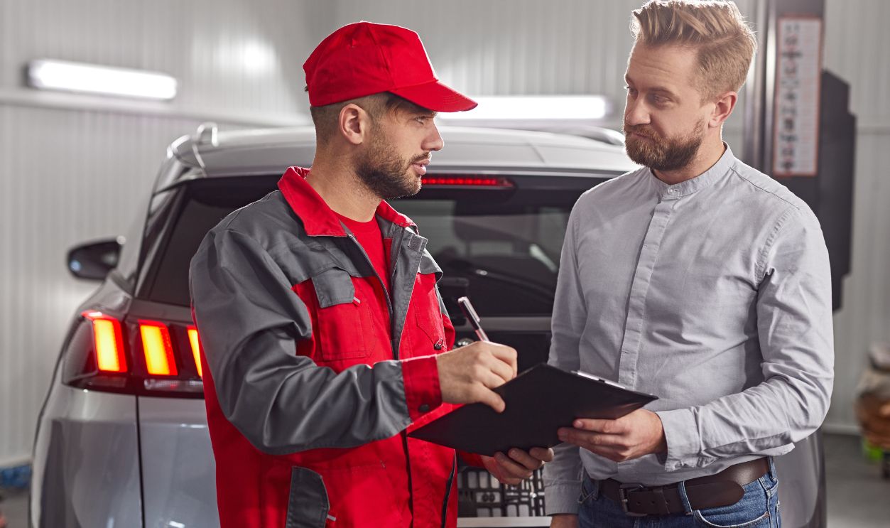 Professional mechanic making notes in checklist document while discussing car repair with male client in modern workshop