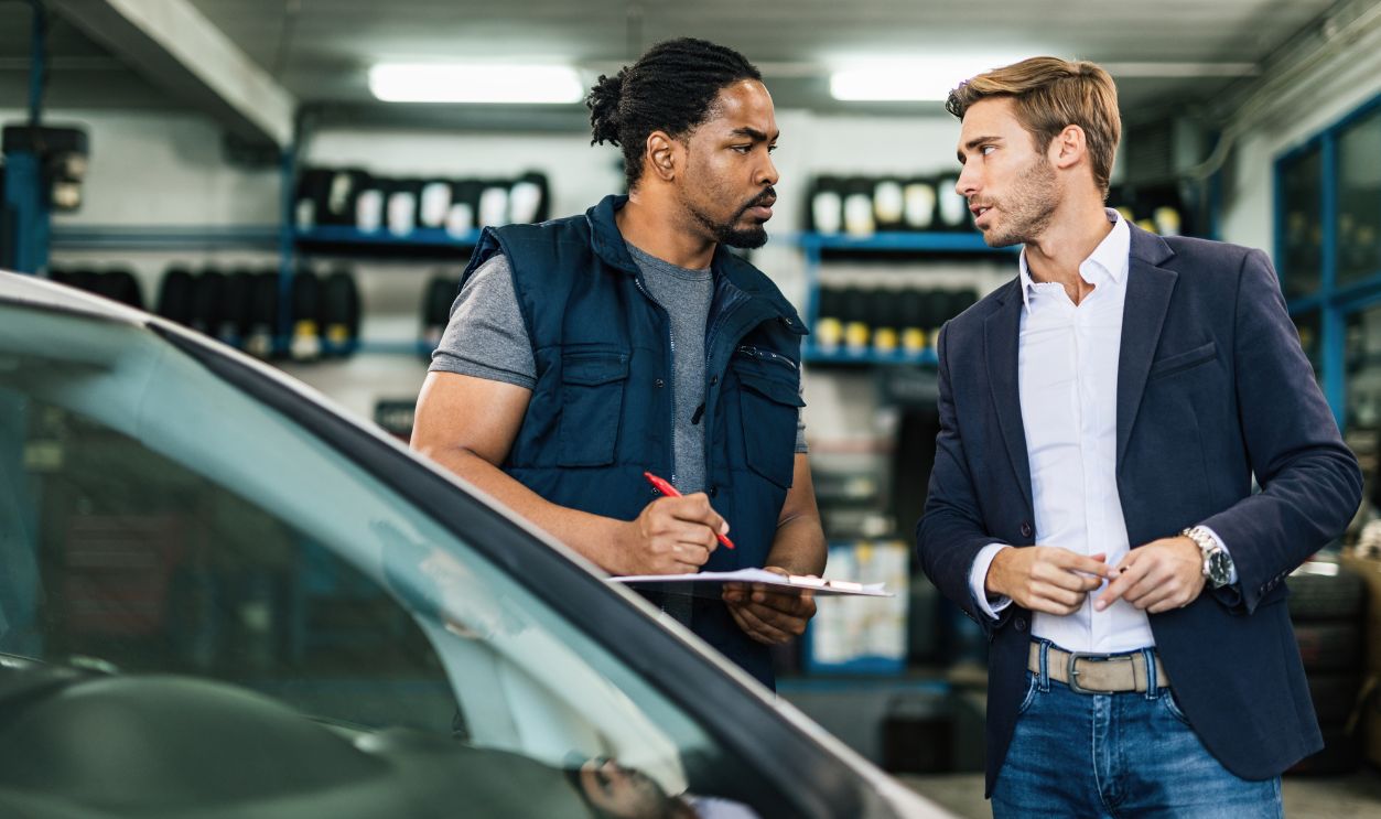 Black car mechanic taking notes while talking to young businessman in auto repair workshop.